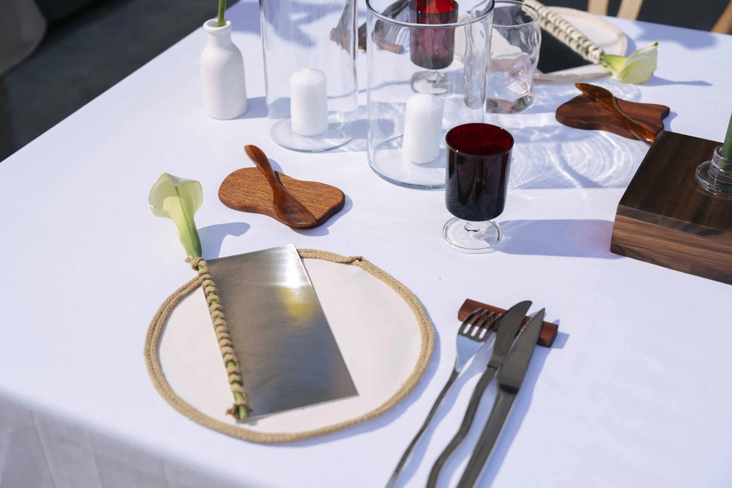 Elegant outdoor dining table set with white tablecloth, cutlery on a wooden rest, wine glasses, a dark glass of wine, small wooden plates with wooden spoons, glass vases with candles, and calla lily flowers.