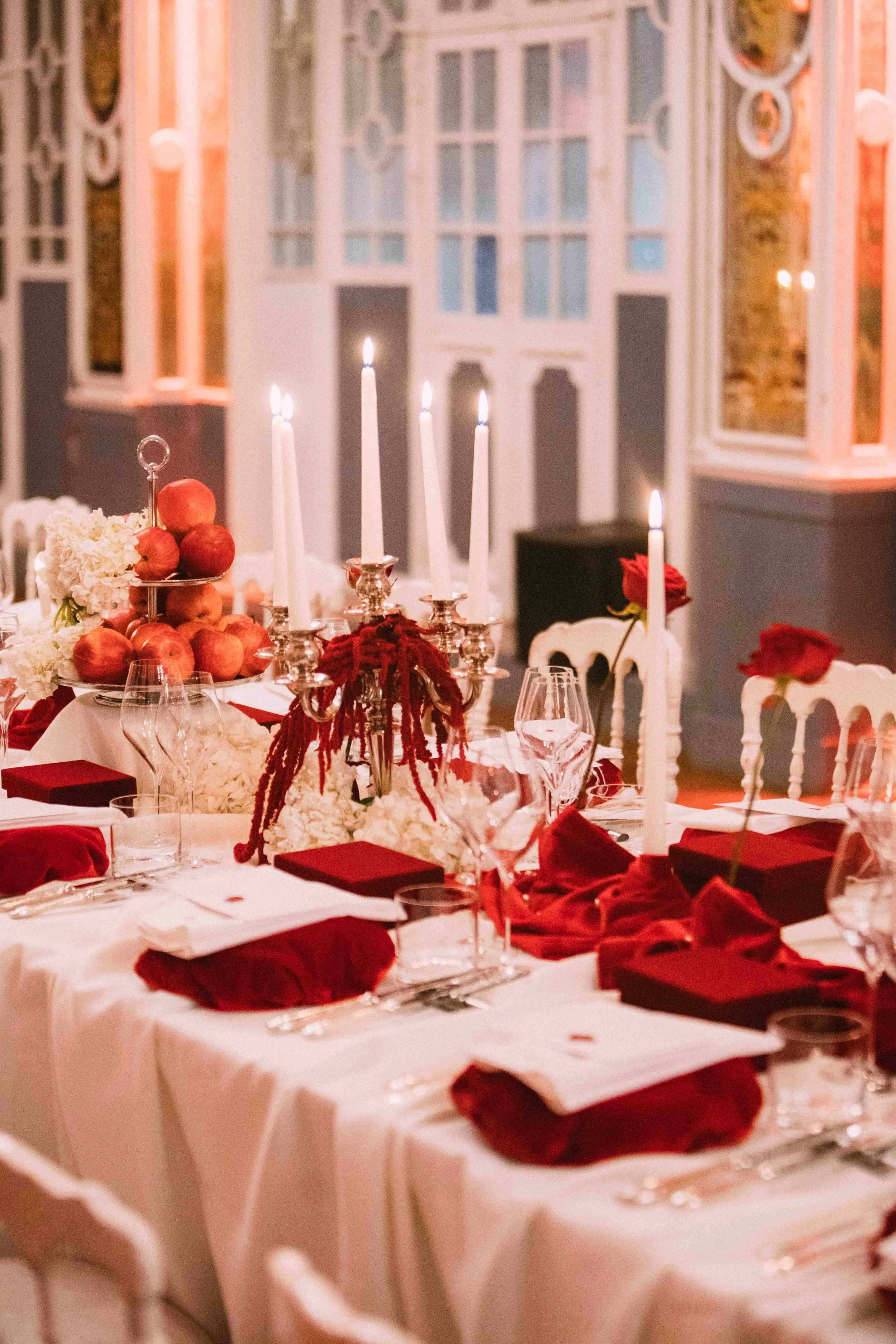 Elegant dining table with white tablecloth, red napkins, wine glasses, candleholders with lit candles, and a centerpiece of white flowers, red roses, and apples, set for a formal event in a decorated room.
