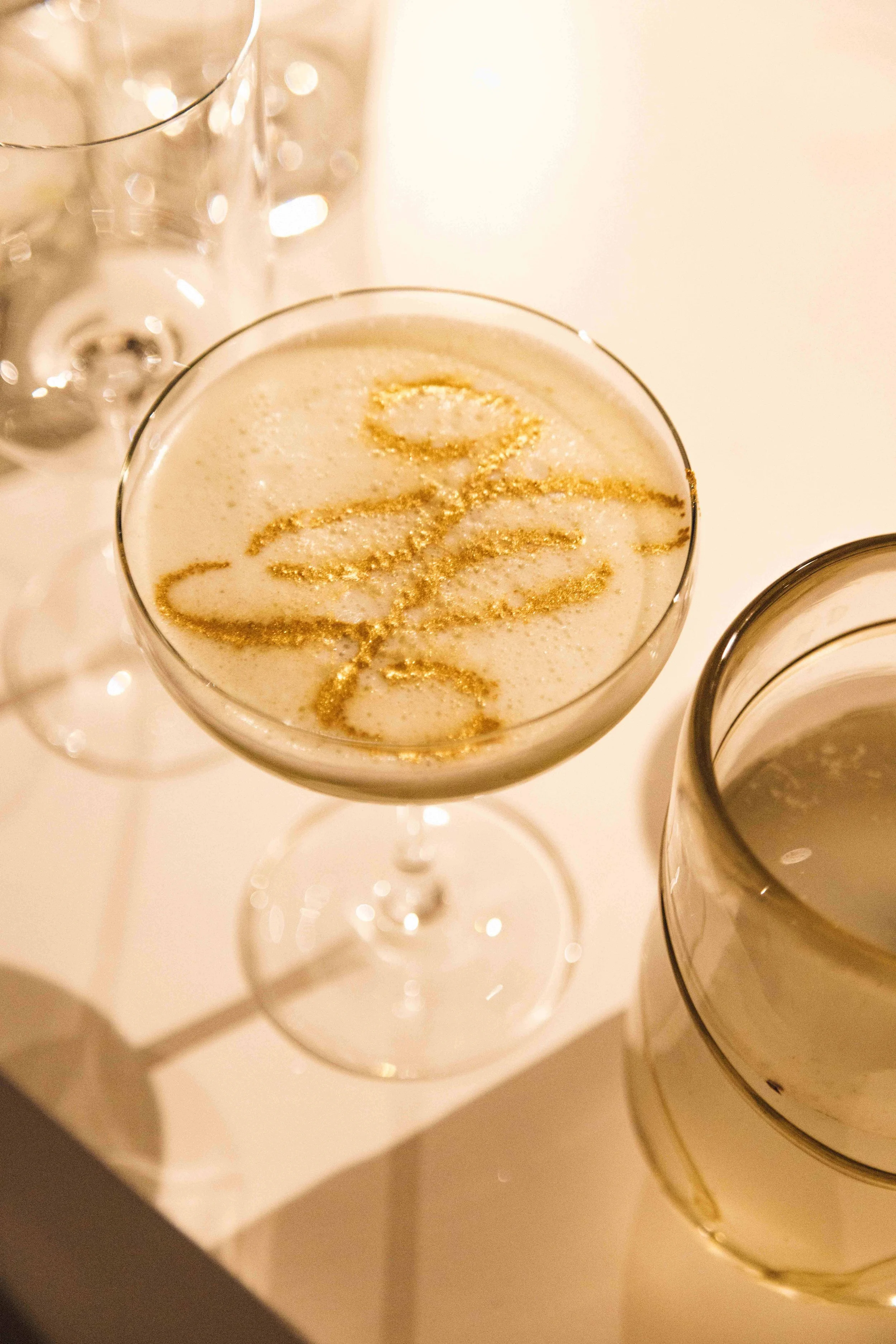 A champagne glass with a frothy beverage decorated with gold glitter that spells out a word, next to a candle and additional glasses.