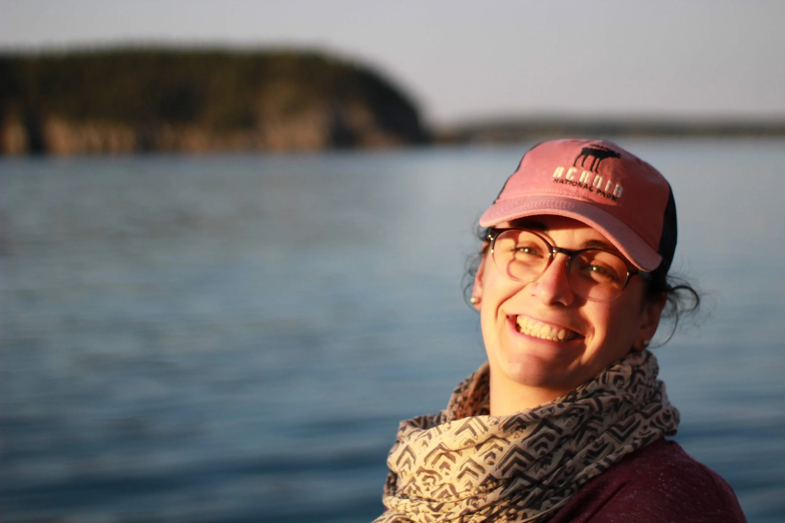 A woman smiling, wearing glasses, a pink cap with the words "ACOEGA NATIONAL PARK," and a patterned scarf, standing near water with a blurred landscape in the background.