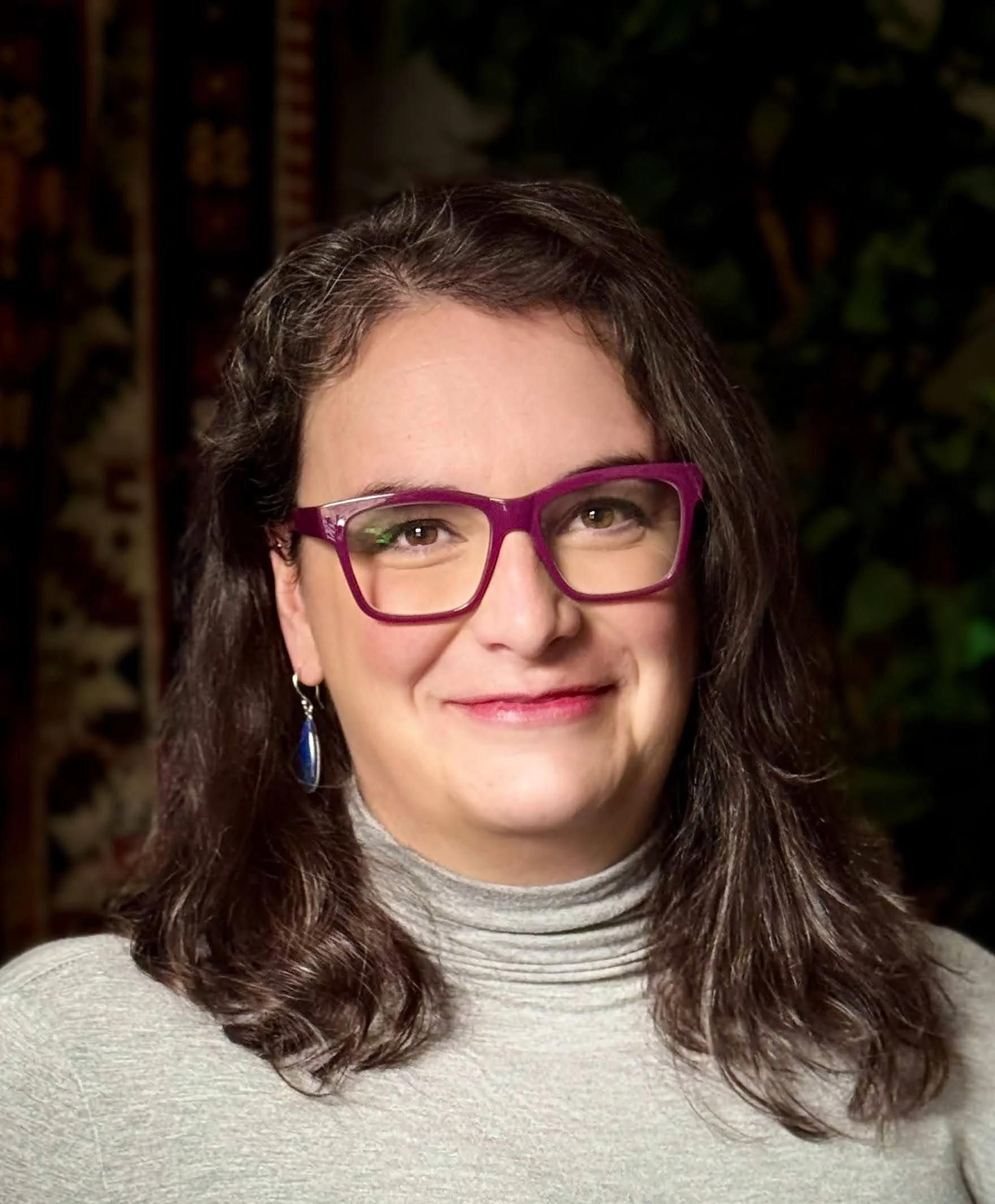 A woman with shoulder-length curly brown hair wearing purple glasses, a gray turtleneck sweater, and blue earrings, smiling in front of a dark, patterned background.