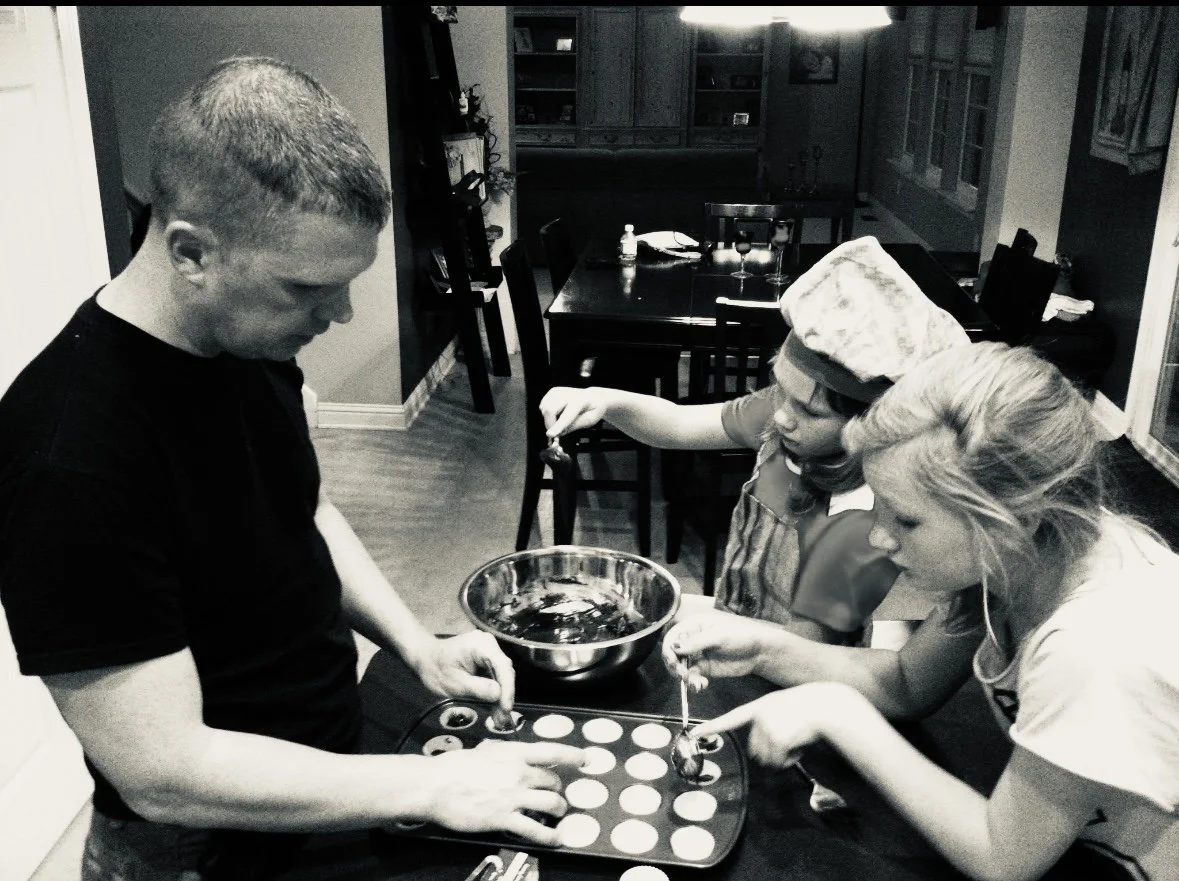 Two women and a man preparing cookies in a kitchen. The man is placing cookie dough on a baking sheet, while one woman is using a spoon to place dough and the other woman is holding a mixing bowl.