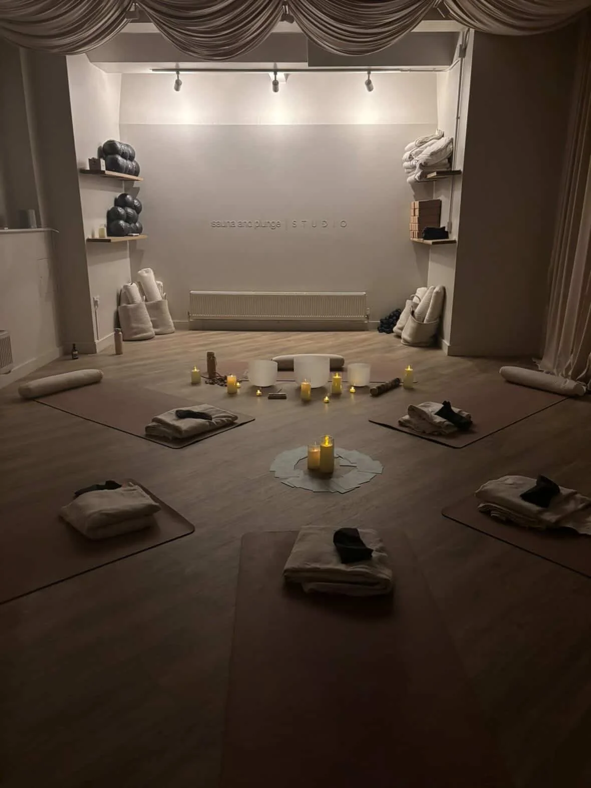 A dimly lit yoga or meditation studio with candles, folded towels, mats, and meditation cushions arranged for a session, with shelves holding robes and meditation props, and a wall with the text 'soul and plunge STUDIO'.