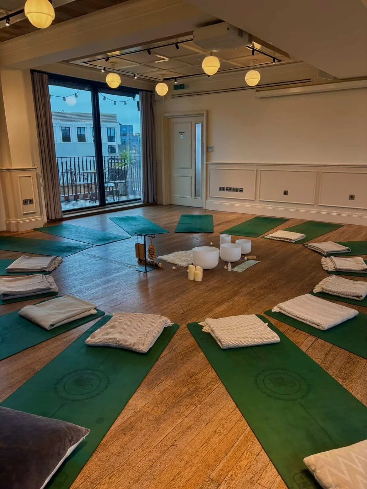 Indoor yoga class setup with green mats and folded towels, a few candles, and singing bowls in a room with wooden floors, large sliding glass door, and balcony outside.