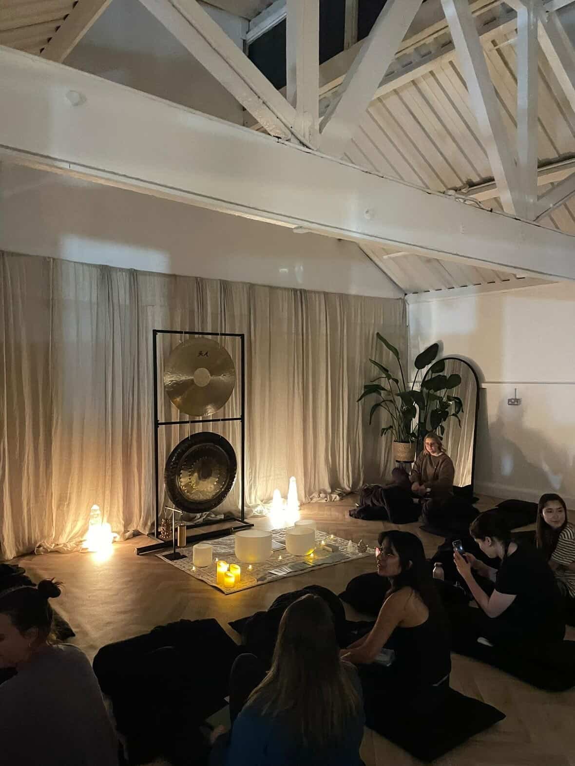 People sitting on yoga mats in a dimly lit room with candles, a large plant, and musical instruments set up for a meditation or yoga session.