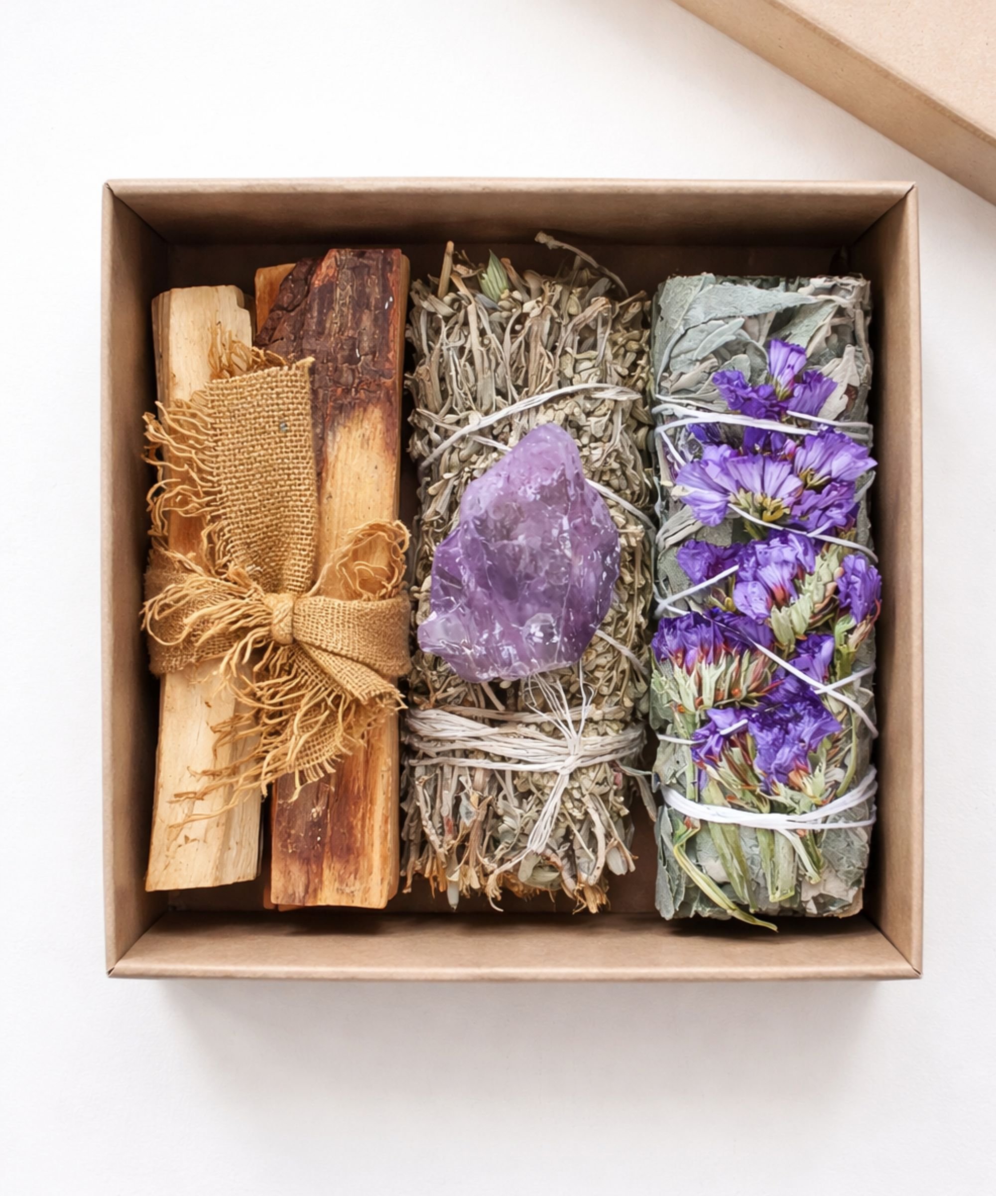 Kraft gift box containing palo santo sticks tied with hessian, a white sage bundle with raw amethyst, and a floral sage bundle with purple blooms.