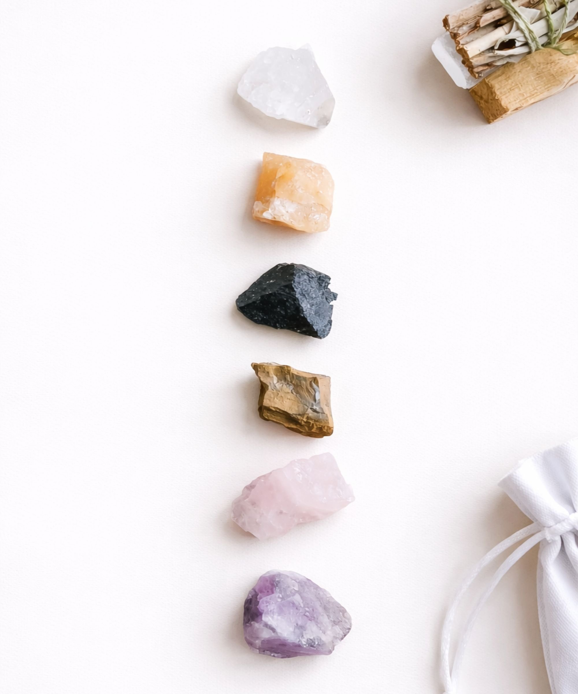 Five raw crystals in a diagonal line — clear quartz, peach calcite, black tourmaline, tiger's eye, rose quartz and amethyst — with a sage bundle and white drawstring pouch alongside.