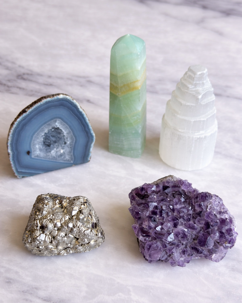 Five crystals on marble: a blue agate slice, green fluorite tower, white selenite, silver pyrite chunk, and a purple amethyst cluster.