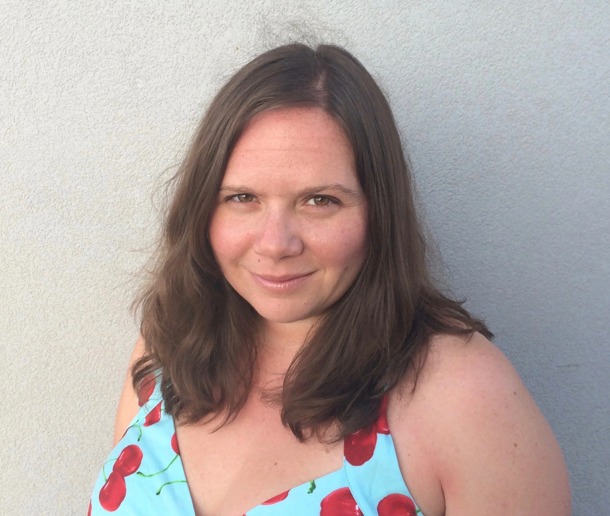 A woman with shoulder-length brown hair, smiling, wearing a sleeveless dress with cherries on a blue background, standing against a light gray wall.
