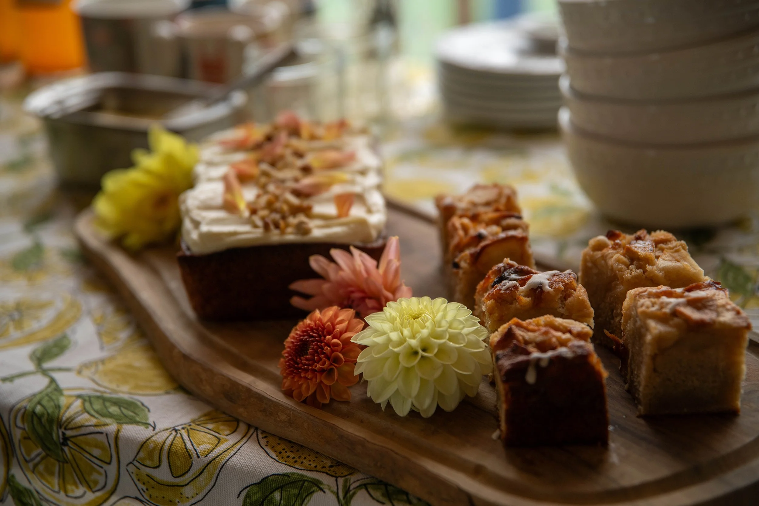 Assorted desserts on a wooden serving tray, decorated with flowers, with a tablecloth featuring a lemon pattern underneath.