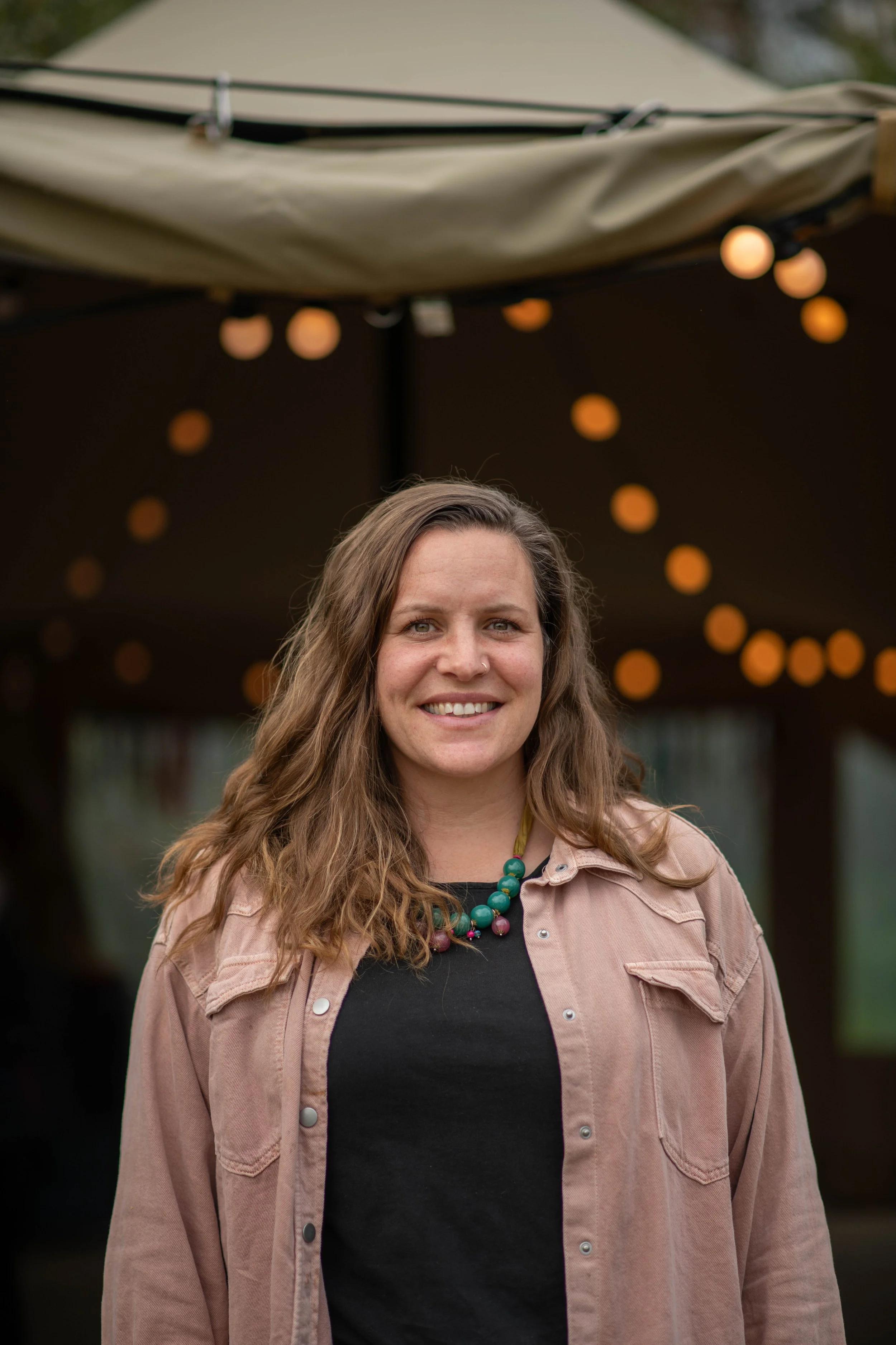 A woman with long wavy brown hair smiling at the camera outside under a stretch tent with festoon lights overhead.