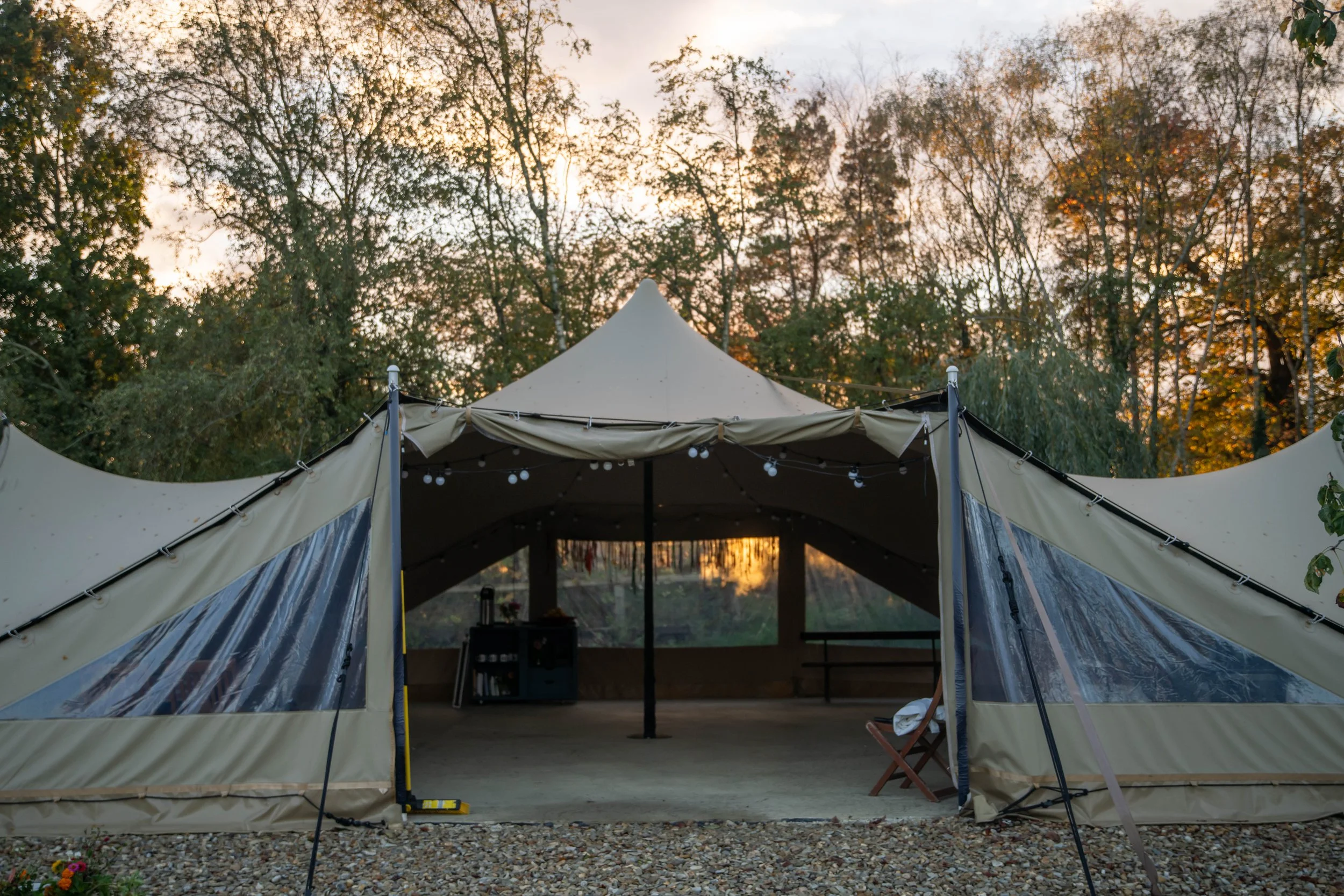 A large outdoor tent set up in a wooded area during sunset, with trees displaying autumn foliage in the background, and string lights hanging inside the tent.