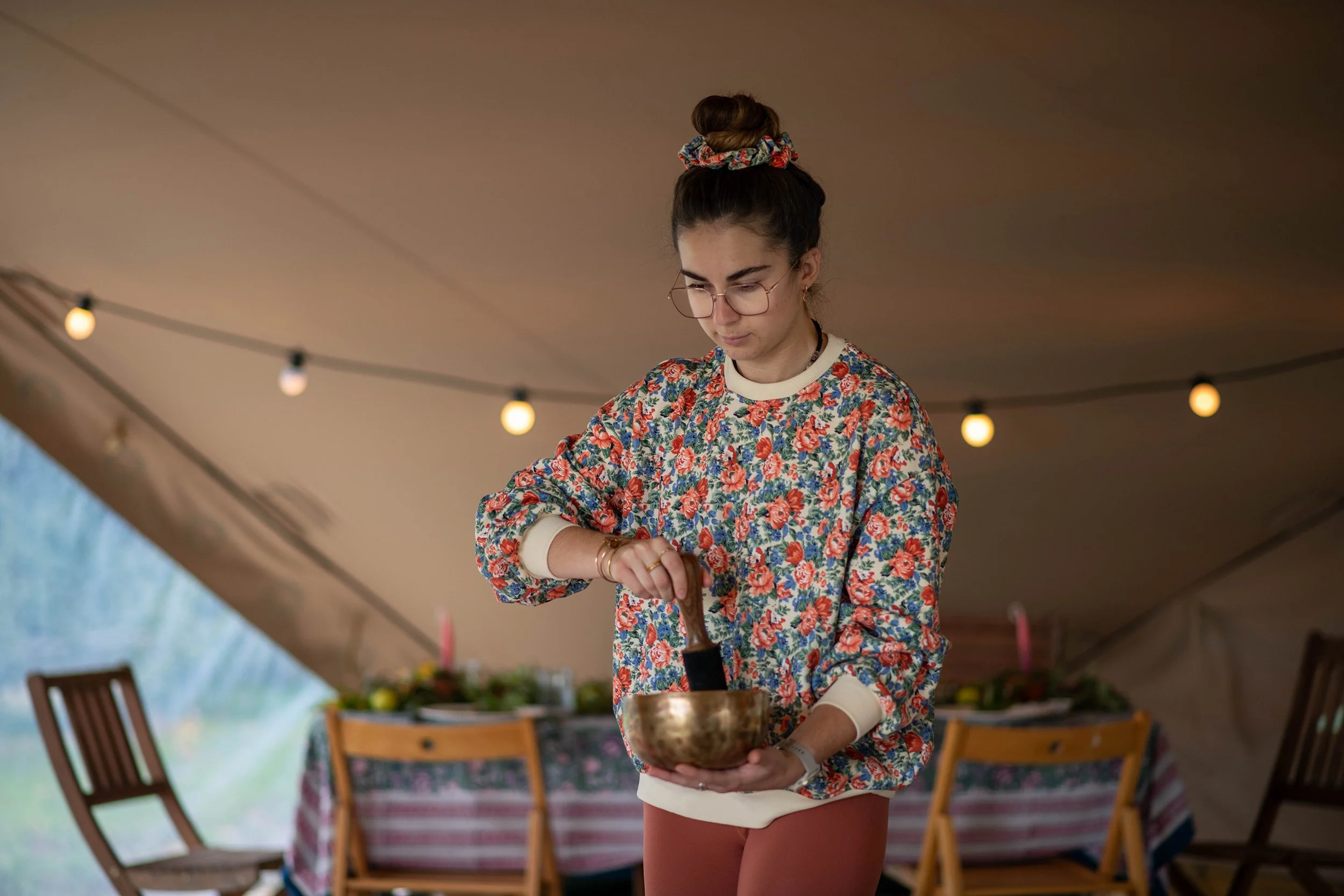 A young woman with glasses and a floral shirt standing under a tent, holding a brass bowl, with a dining table and chairs in the background.