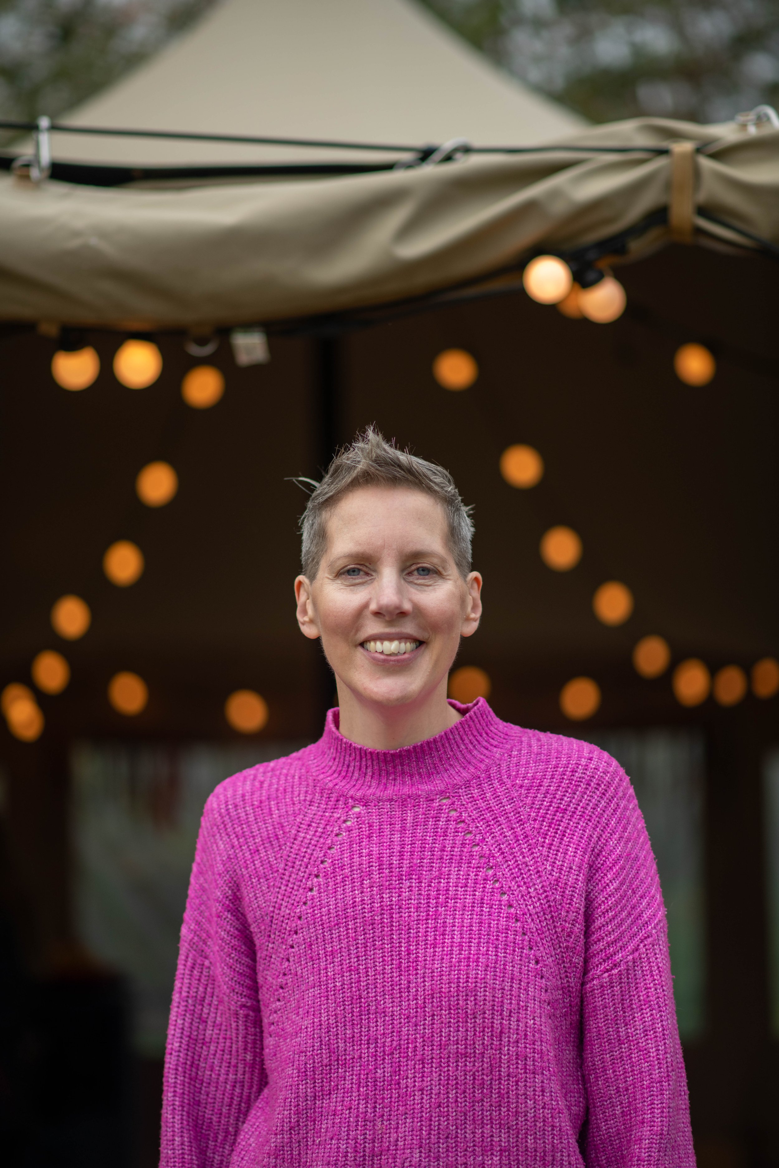 A woman smiling in front of a string of hanging warm-toned lights at an outdoor event, wearing a bright pink sweater.