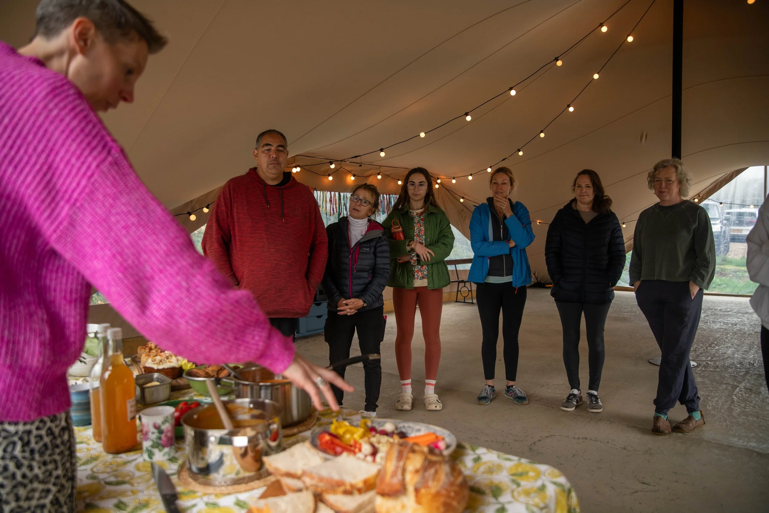 A woman in a pink sweater with leopard print pants is serving food, including bread, sliced vegetables, and condiments, at a table set up with various dishes and bottles. In the background, six people in casual outdoor clothing are watching her inside a large tent decorated with string lights.