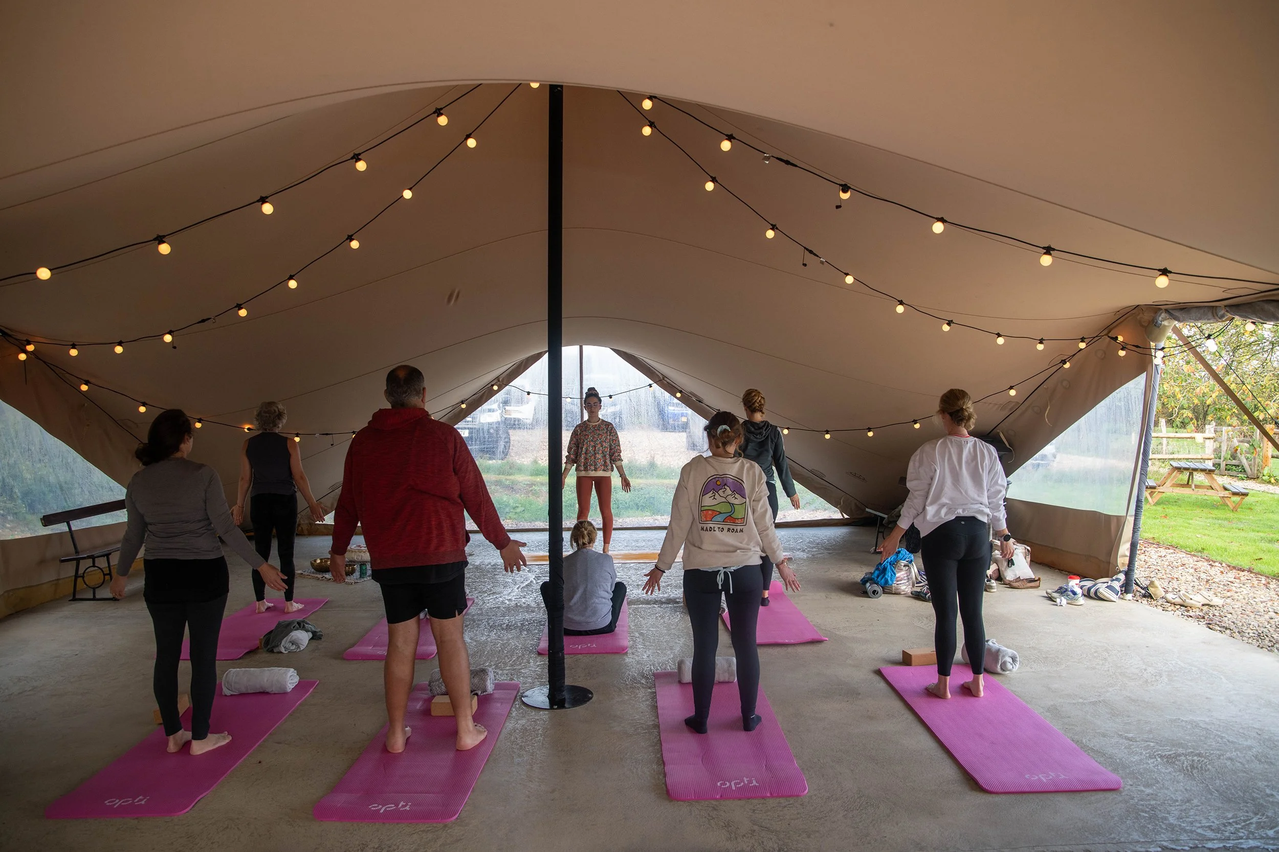 People participating in a yoga class inside a large tent with string lights hanging overhead, yoga mats on the floor, some with rolled towels, during daytime with outdoor scenery visible through the open side of the tent.