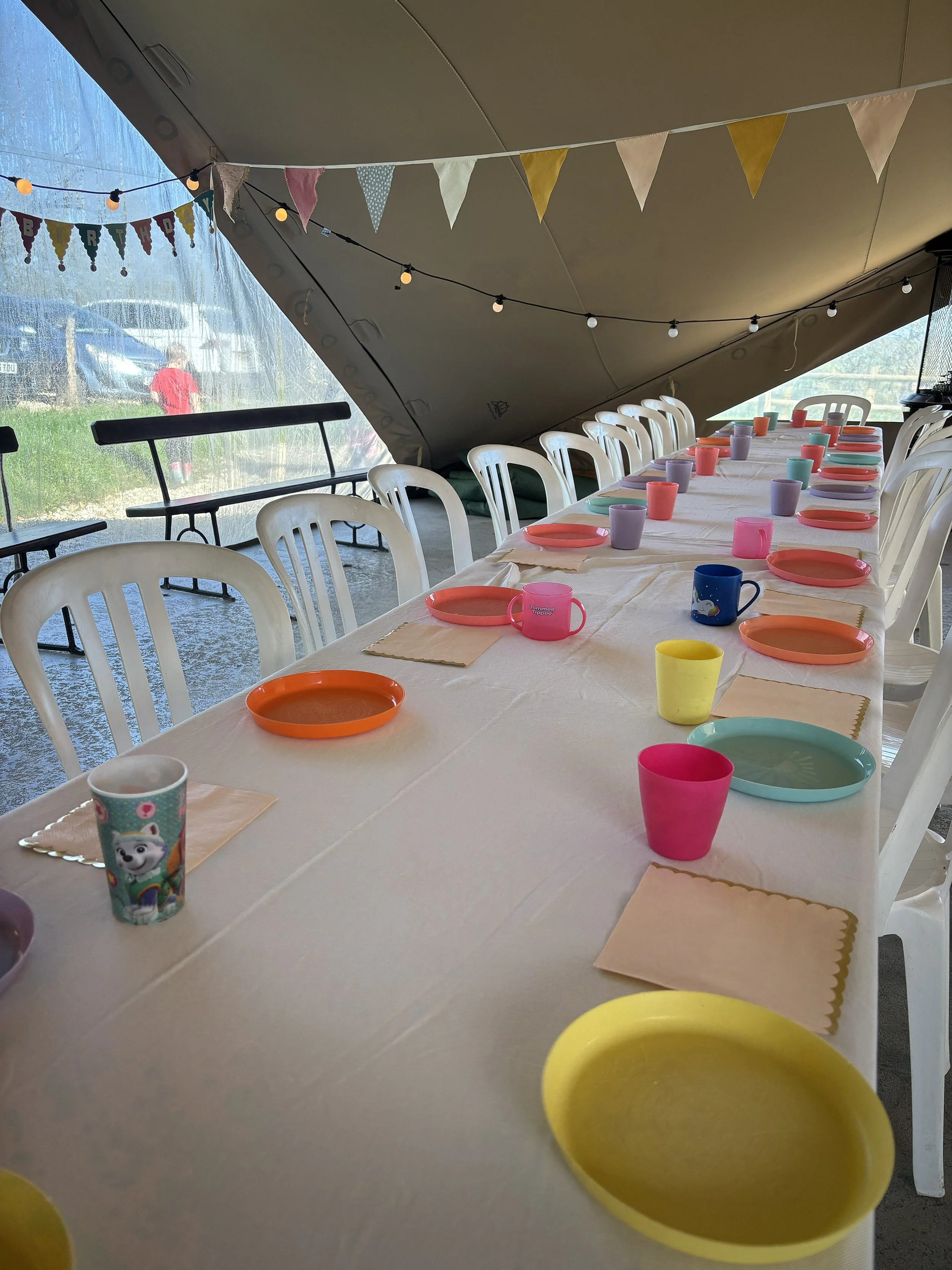 Decorated long table with colorful paper plates and cups, set for a children's birthday party inside a stretch tent with bunting and string lights.