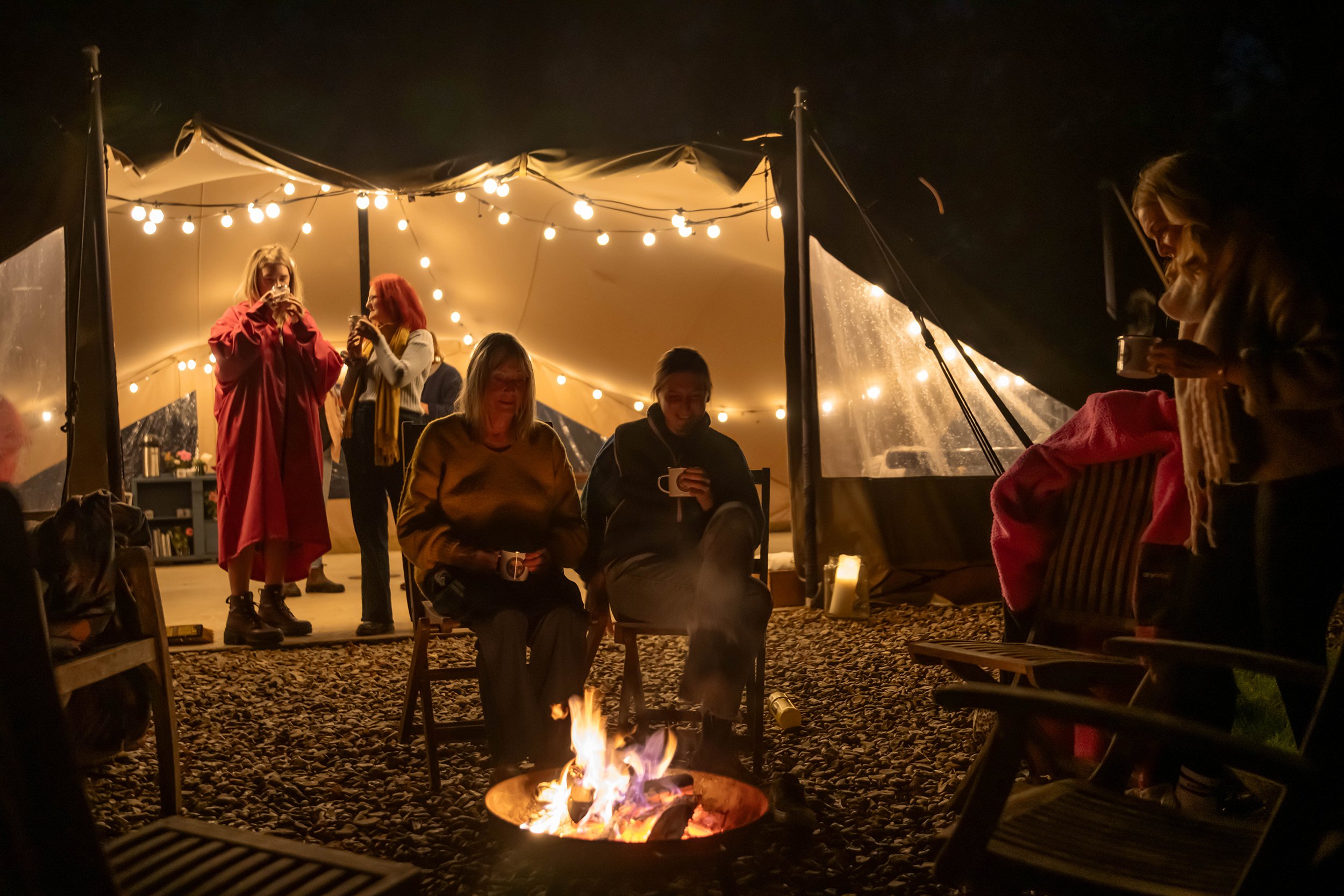 Group of friends enjoying a cozy evening around a campfire under string lights, with some sitting and drinking, others standing and chatting, in a tented outdoor setting at night.