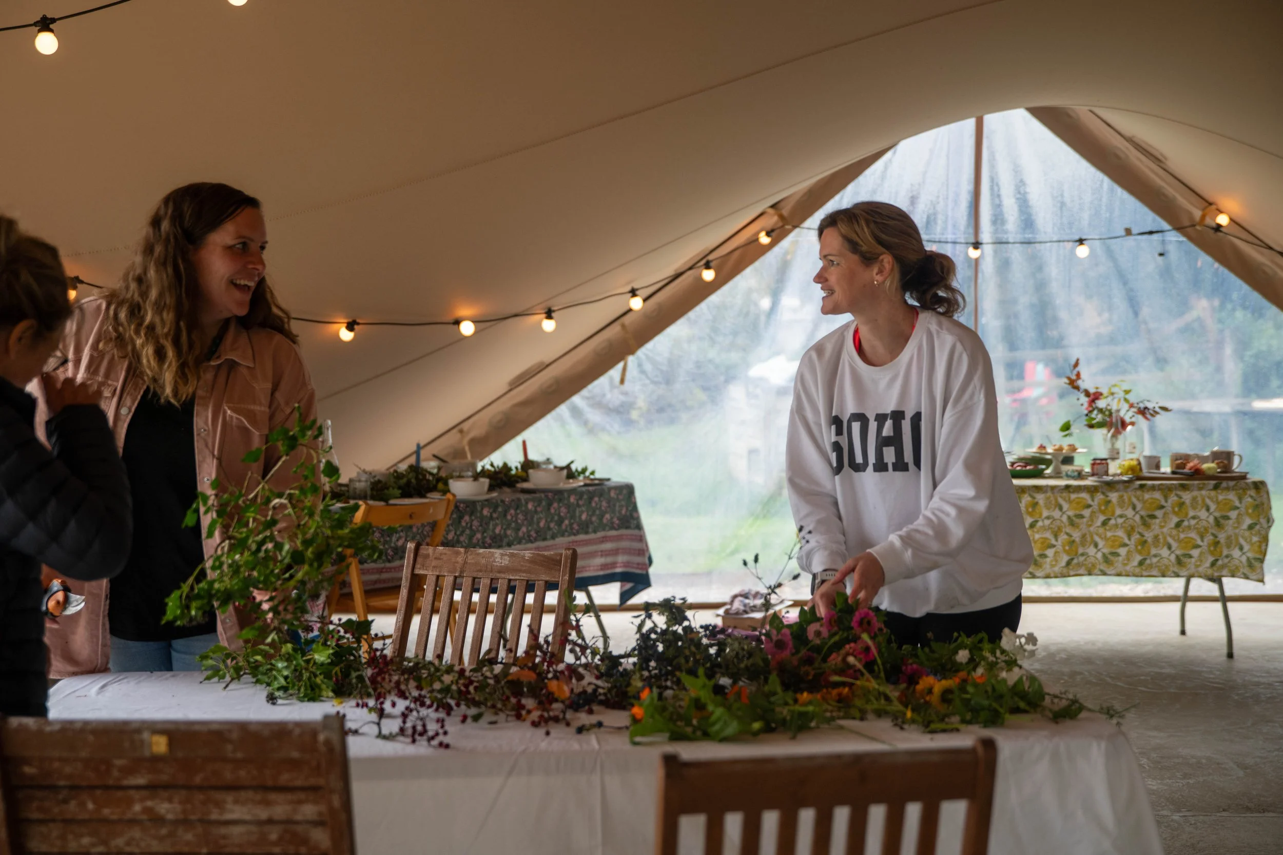 Three women inside a large tent with string lights, making flower arrangements at a table covered with flowers and greenery, with tables in the background set with dishes and decorations.