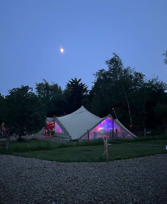 A white tent with colorful lights inside, set up outdoors on grass with trees in the background under a blue sky with the moon visible.