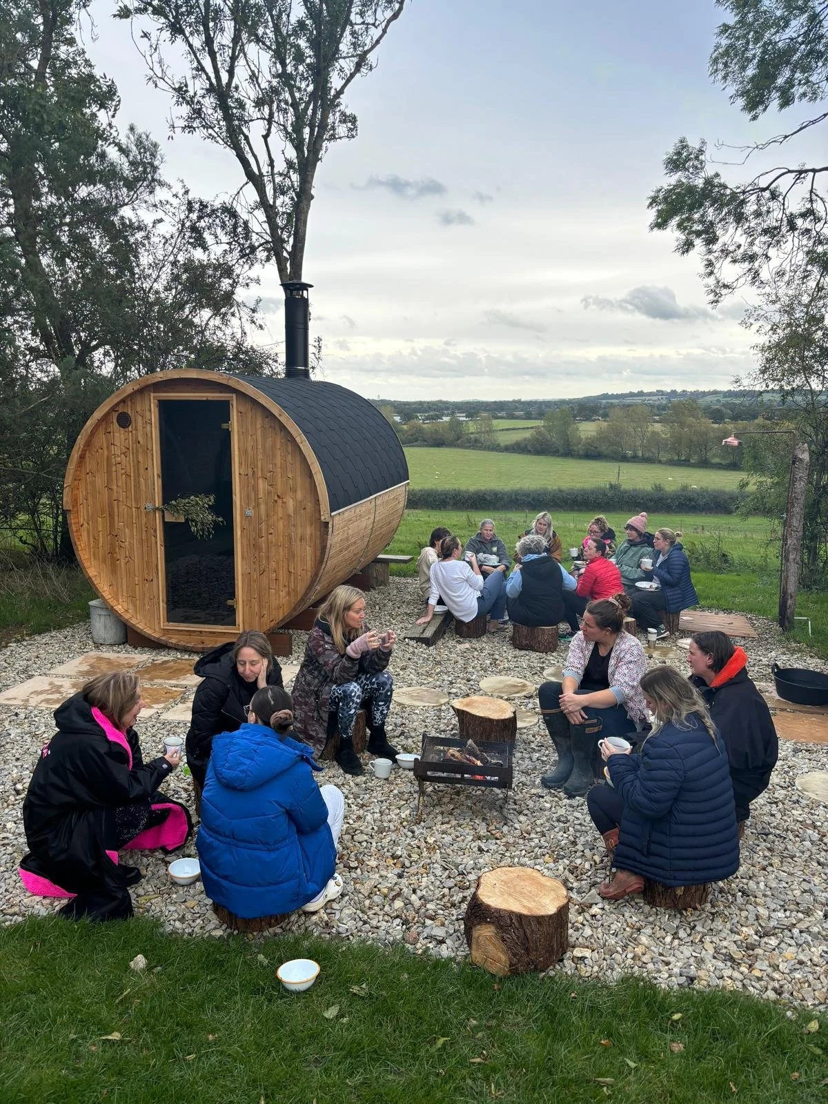 Group of people sitting outside near a wooden sauna on a gravel area, enjoying drinks around a small fire pit, in a rural landscape with trees and fields in the background.