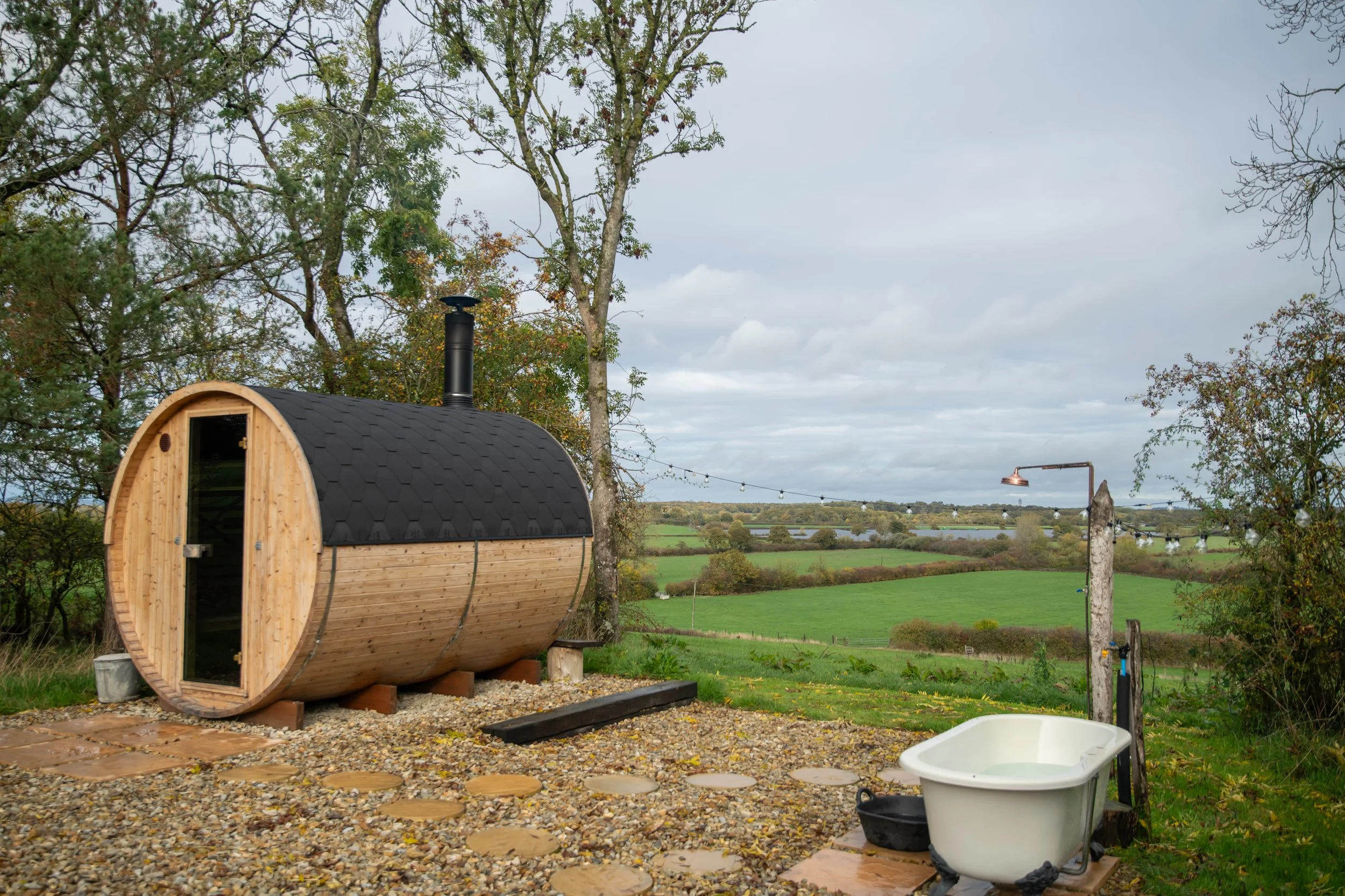 A small wooden sauna with a rounded shape, black shingled roof, located outdoors on a gravel surface, with trees and a landscape of rolling green fields and a river in the background, under a cloudy sky.