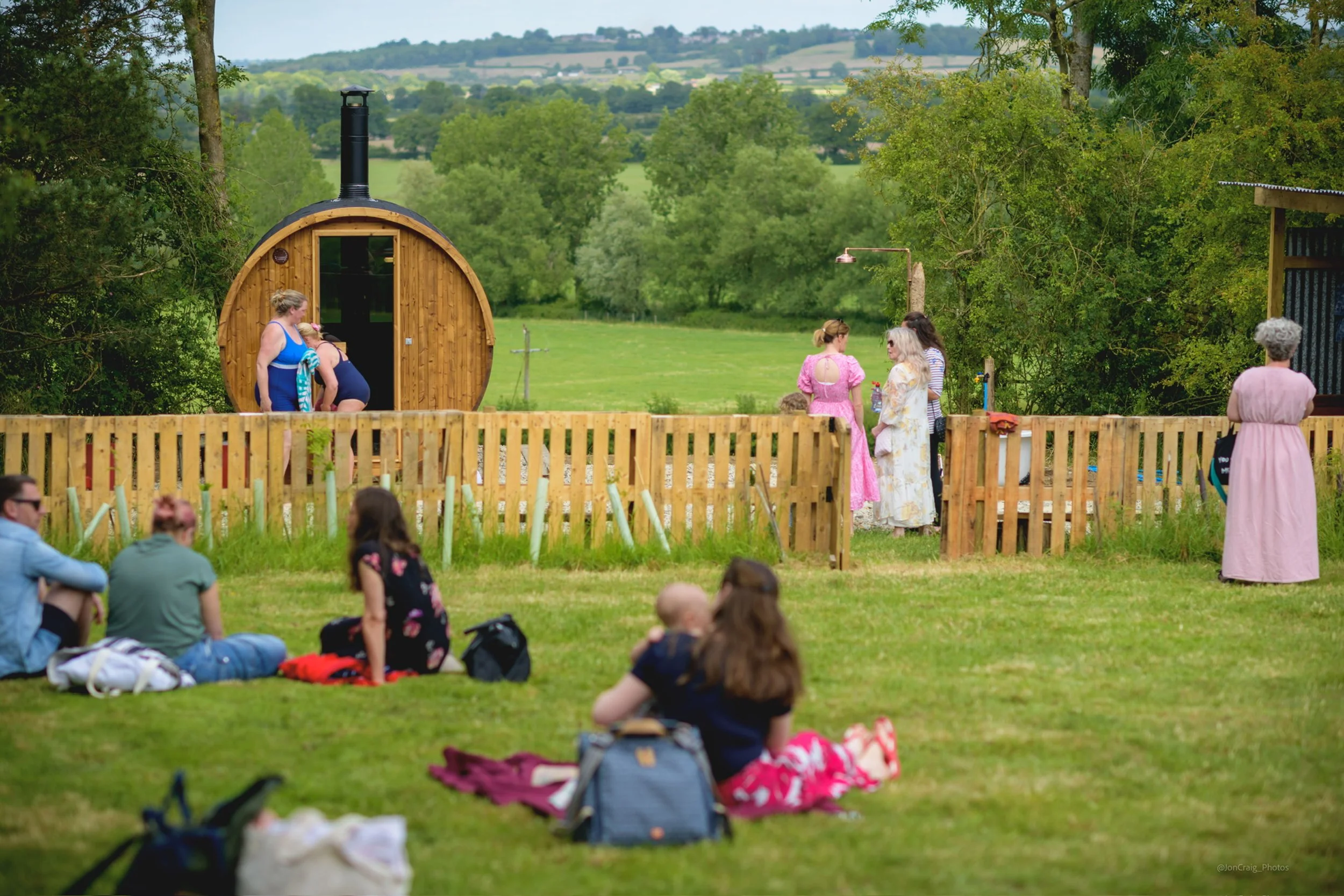 People gather outdoors on a grassy field with a wooden sauna in the background, surrounded by trees and hills, during a sunny day.