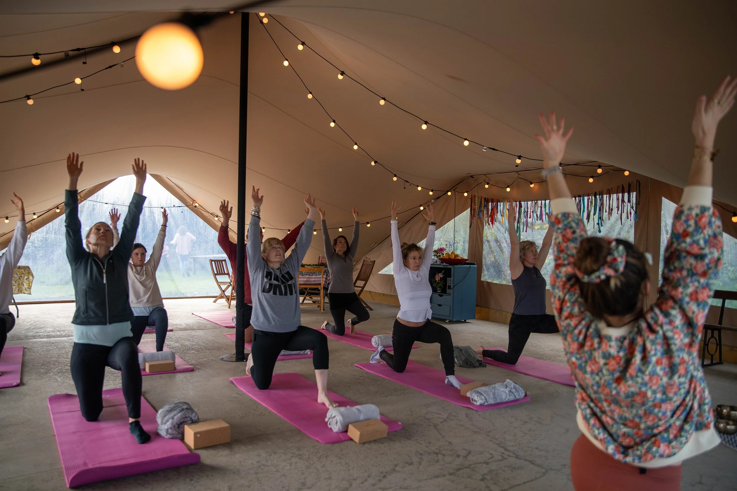 Group of women participating in a yoga class inside a tent, practicing a pose with raised arms on pink yoga mats, surrounded by string lights and decorative colorful ribbons.