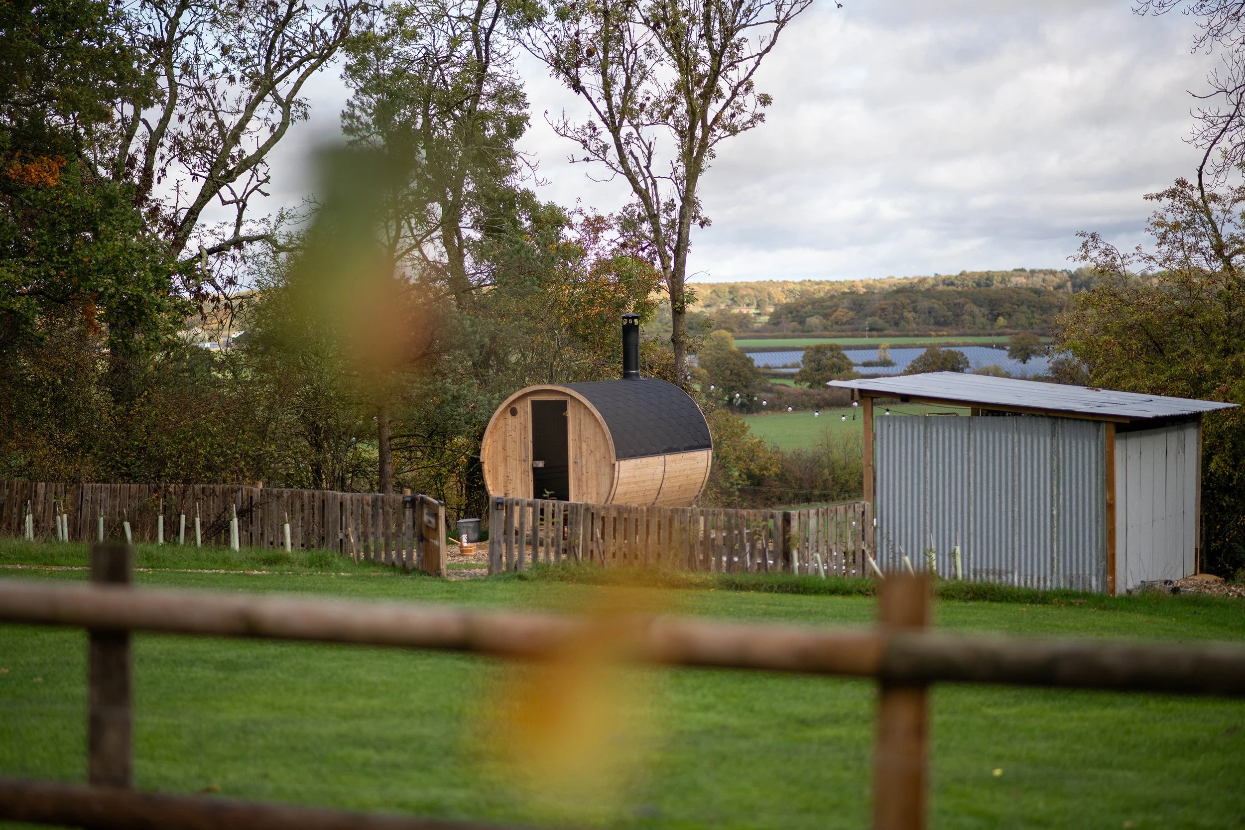 Rural landscape with trees, a small wooden sauna, a metal shed, a fenced yard, and a farm in the background under cloudy sky.