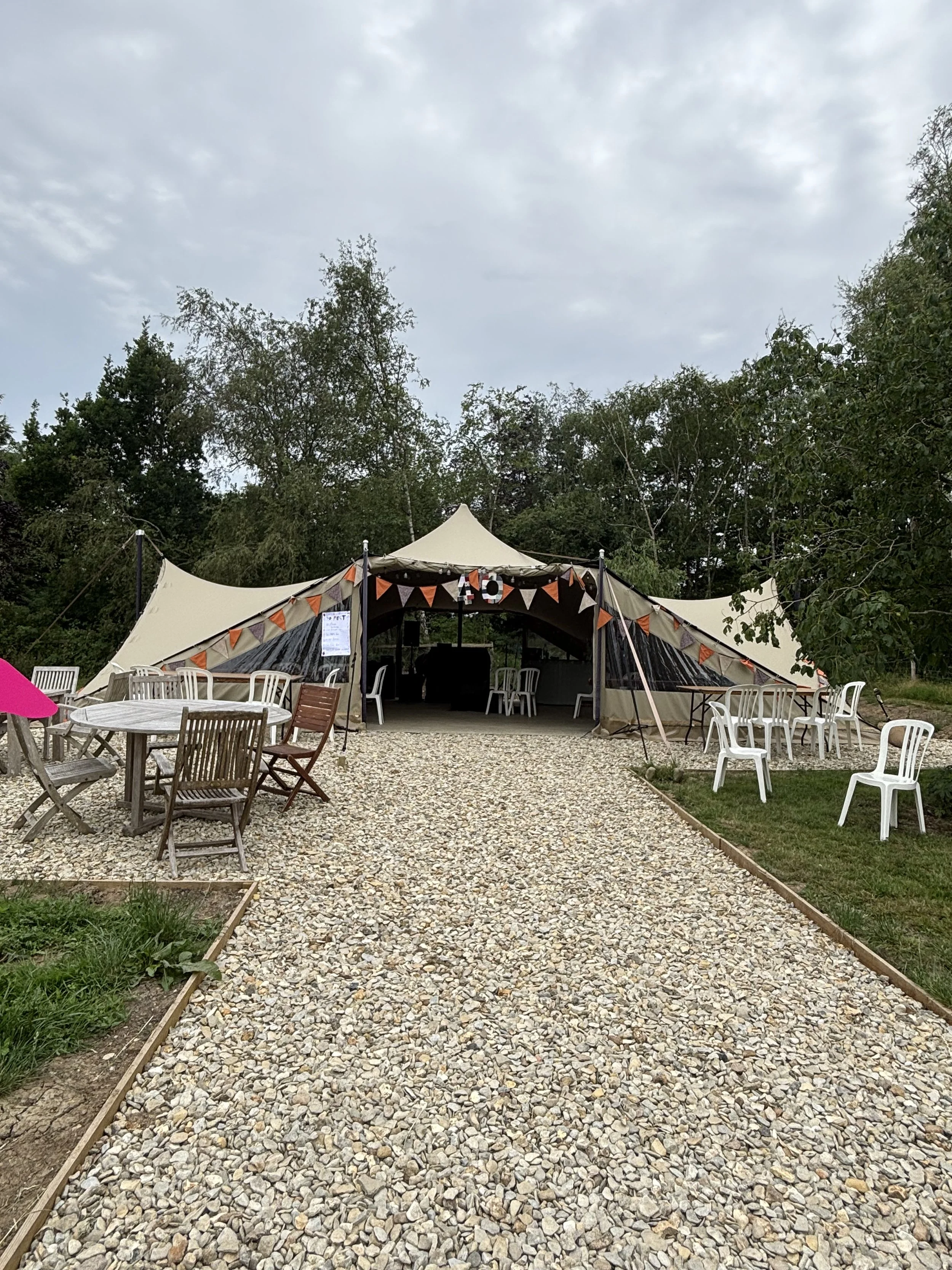 Outdoor event setup with a stretch tent decorated with orange and black bunting, surrounded by white plastic chairs and a wooden table with chairs on a gravel pathway, with tree in the background.
