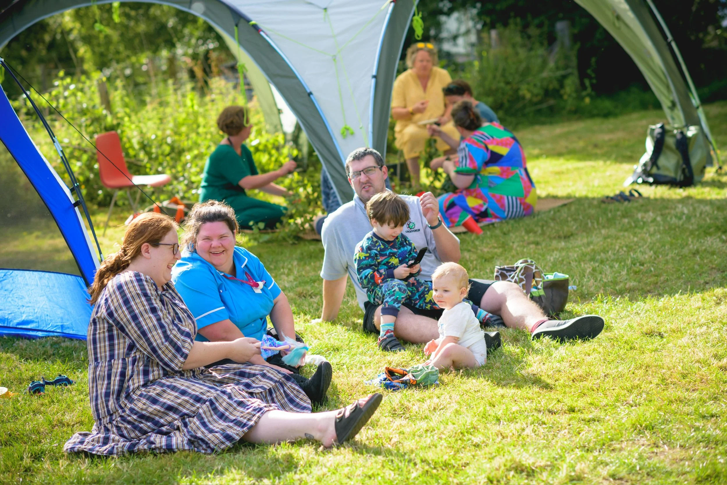 Group of people enjoying a picnic outside, sitting on grass under a camping tent on a sunny day.