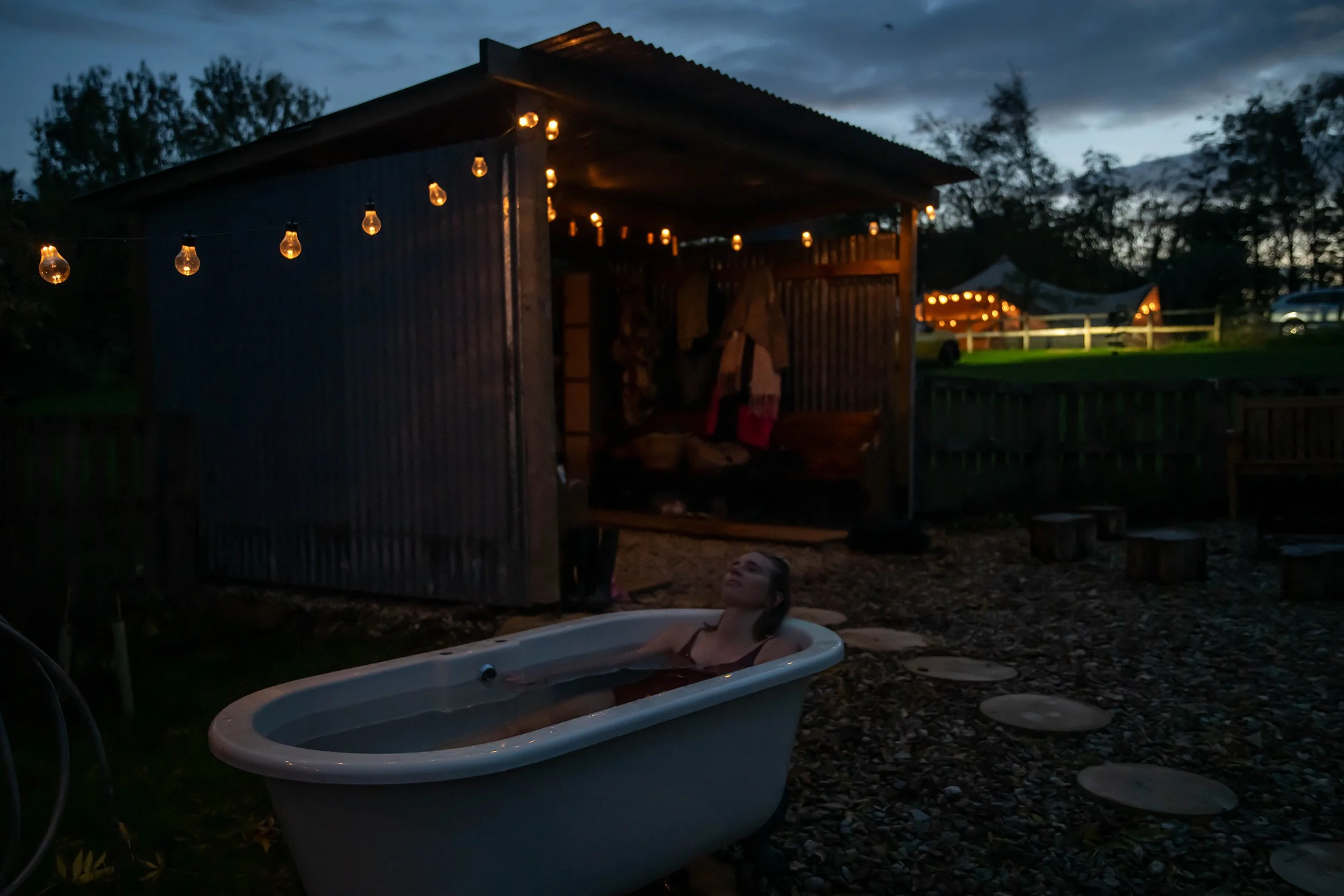 A woman relaxing in a white bathtub outside during evening, with string lights hanging from a wooden shed behind her. The shed contains hanging clothes, and there are trees, a wooden fence, and a lit tent in the background.