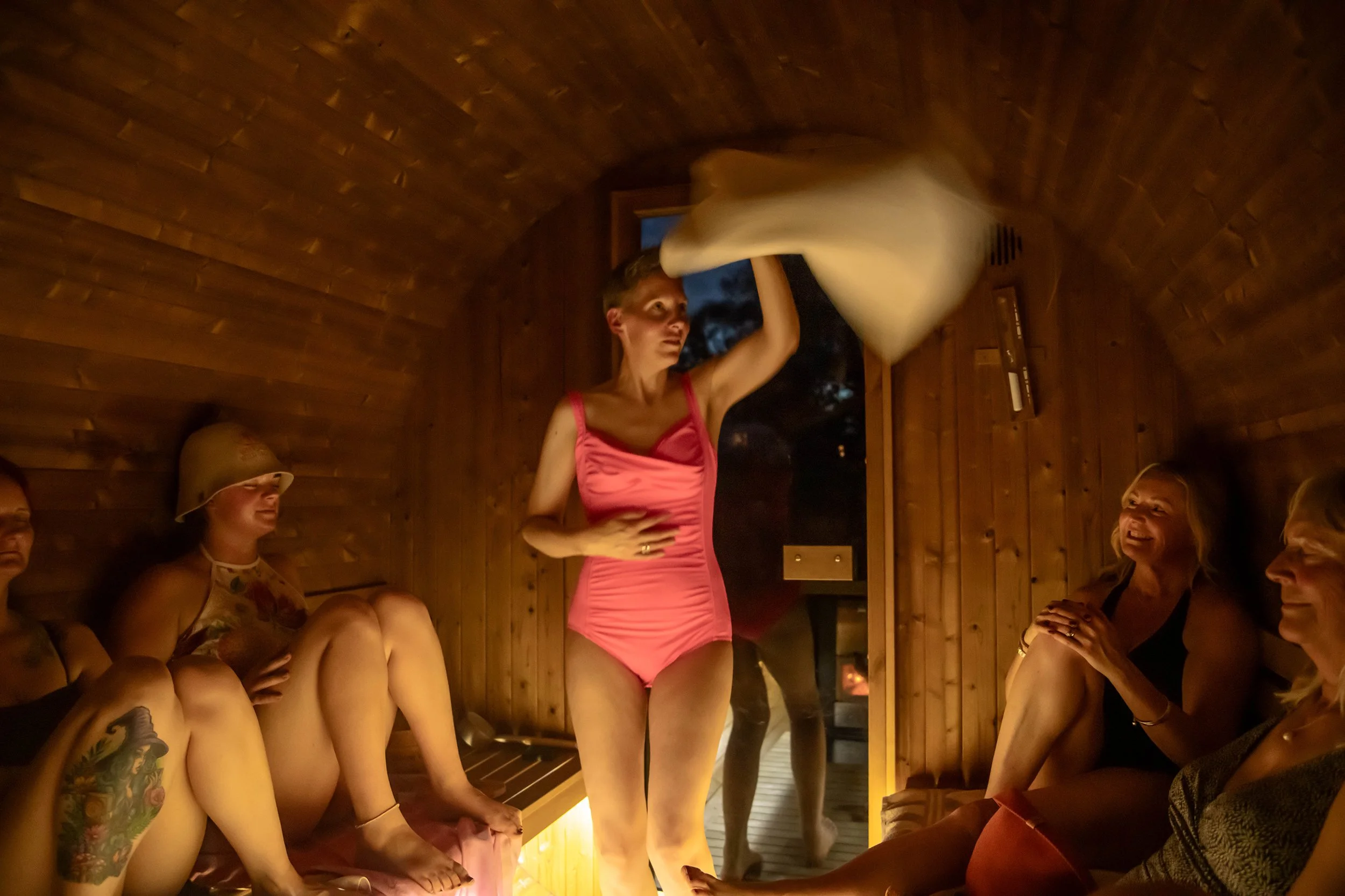 Women enjoying a sauna, some wearing bikinis, with wooden interior walls, and a small window showing the night outside, in a dimly lit setting.