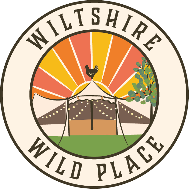 Illustration of a circular emblem with the text 'Wiltshire Wild Place' around the border. Inside, there is a circus tent with a chicken on top, a sunburst background, and a tree on the right side.
