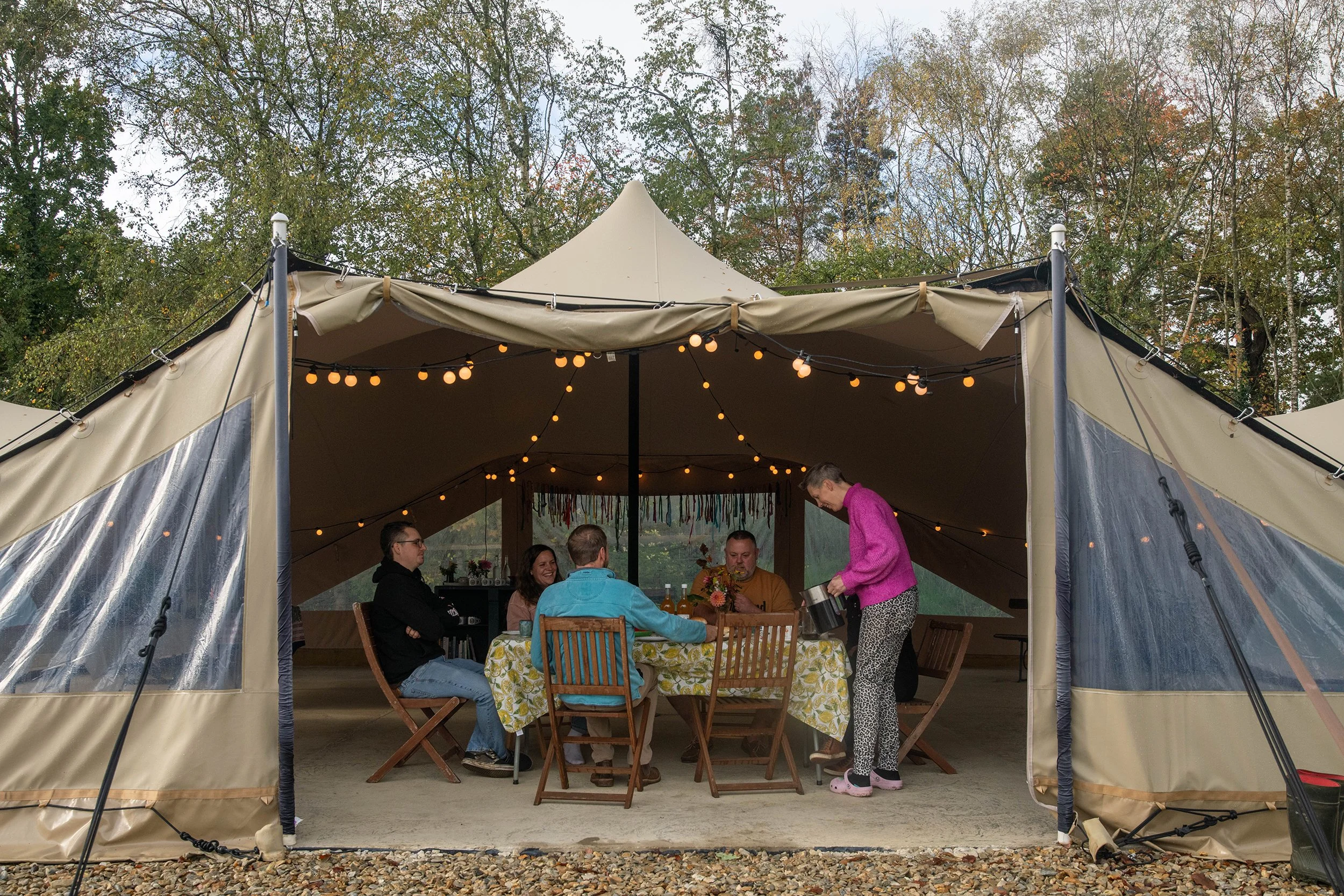 People gathering inside a large outdoor tent with string lights, sitting around a table, with trees in the background.