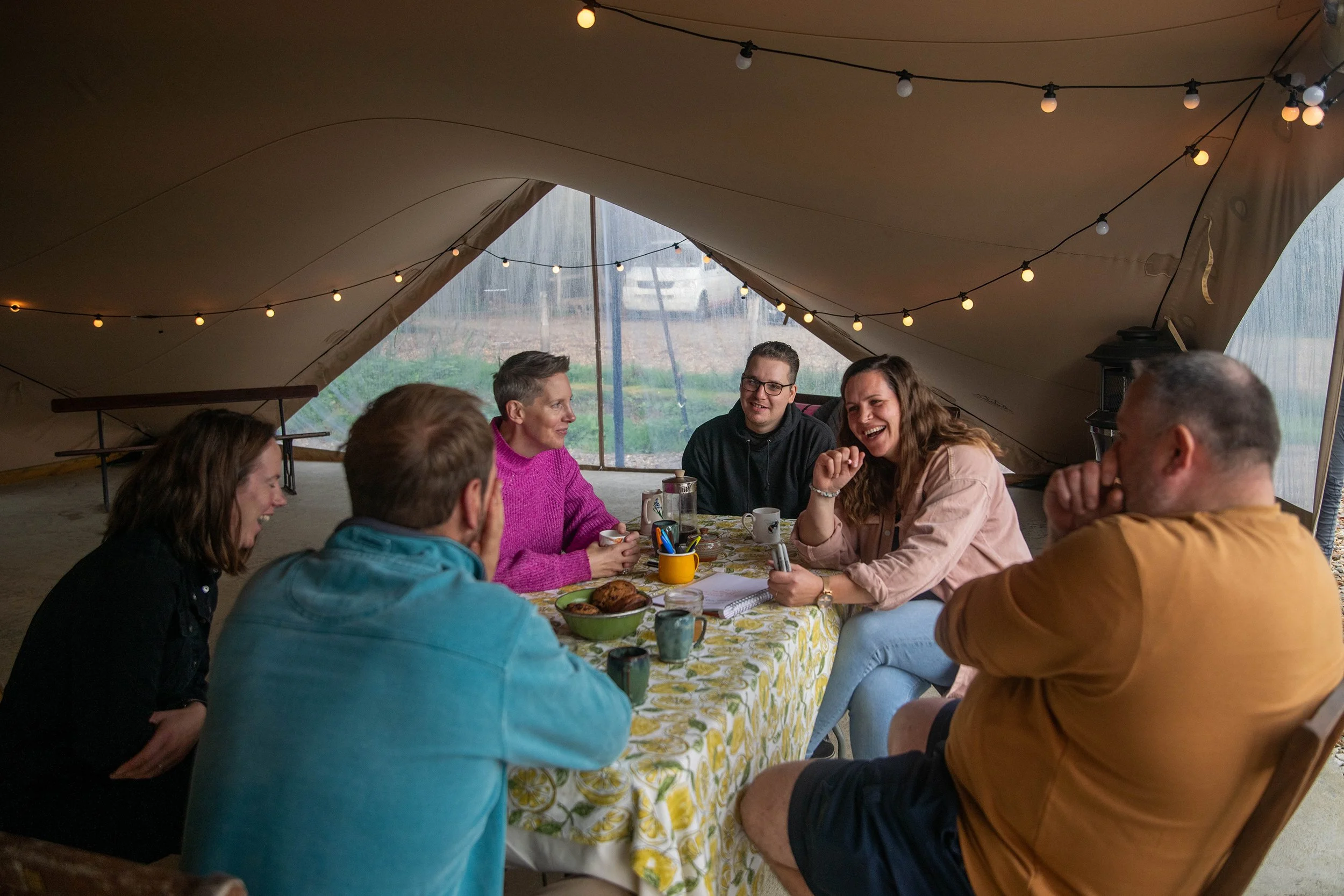 Group of six people sitting around a table in a tent, enjoying a meal and conversation, with string lights hanging above.