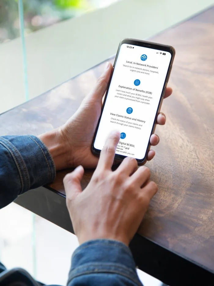 Person using a smartphone with a mental health insurance app open on the screen, sitting at a wooden table near a window.