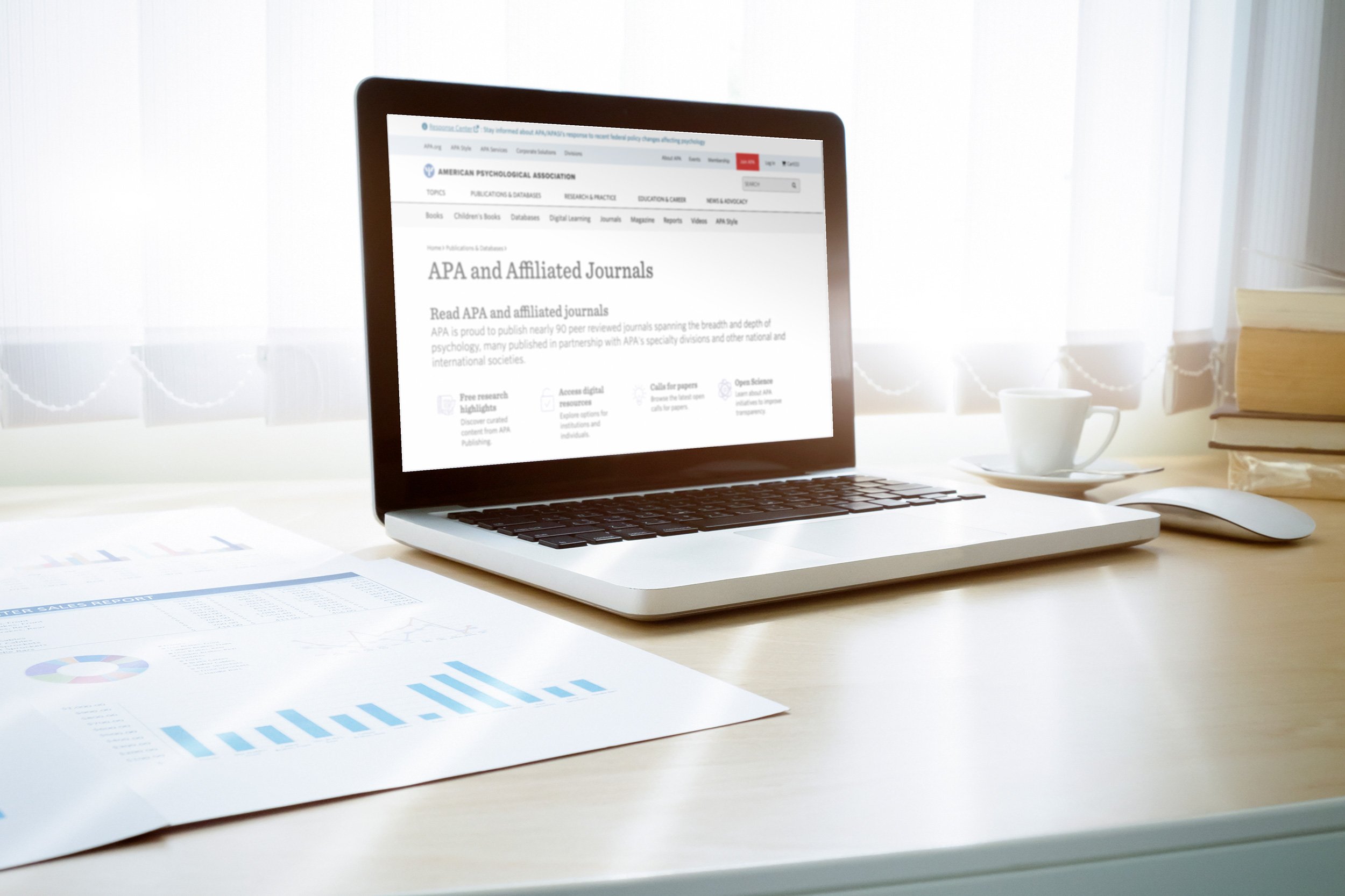 Laptop displaying a webpage for the American Psychological Association, with reports and research articles, surrounded by printed data charts and graphs on a desk.