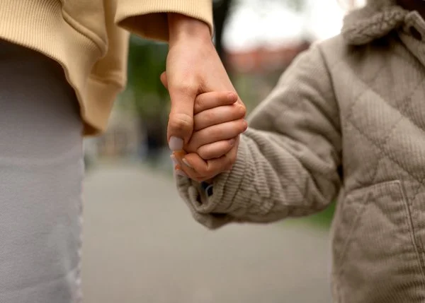 A close-up of an adult and a child holding hands outdoors, with the adult's hand gently clasped around the child's hand.