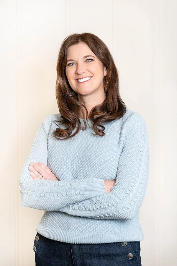 Dr. Berg stands smiling with arms crossed against a light background. She has long brown hair and is wearing a light blue sweater and dark pants.
