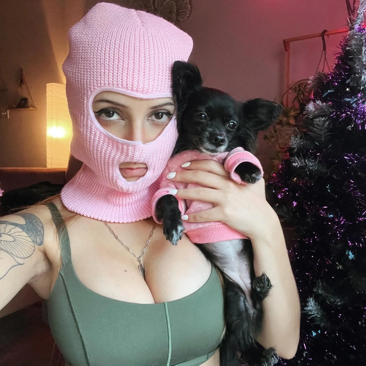 Woman wearing a pink knit ski mask holding a small black and tan dog in a pink sweater, in front of a Christmas tree