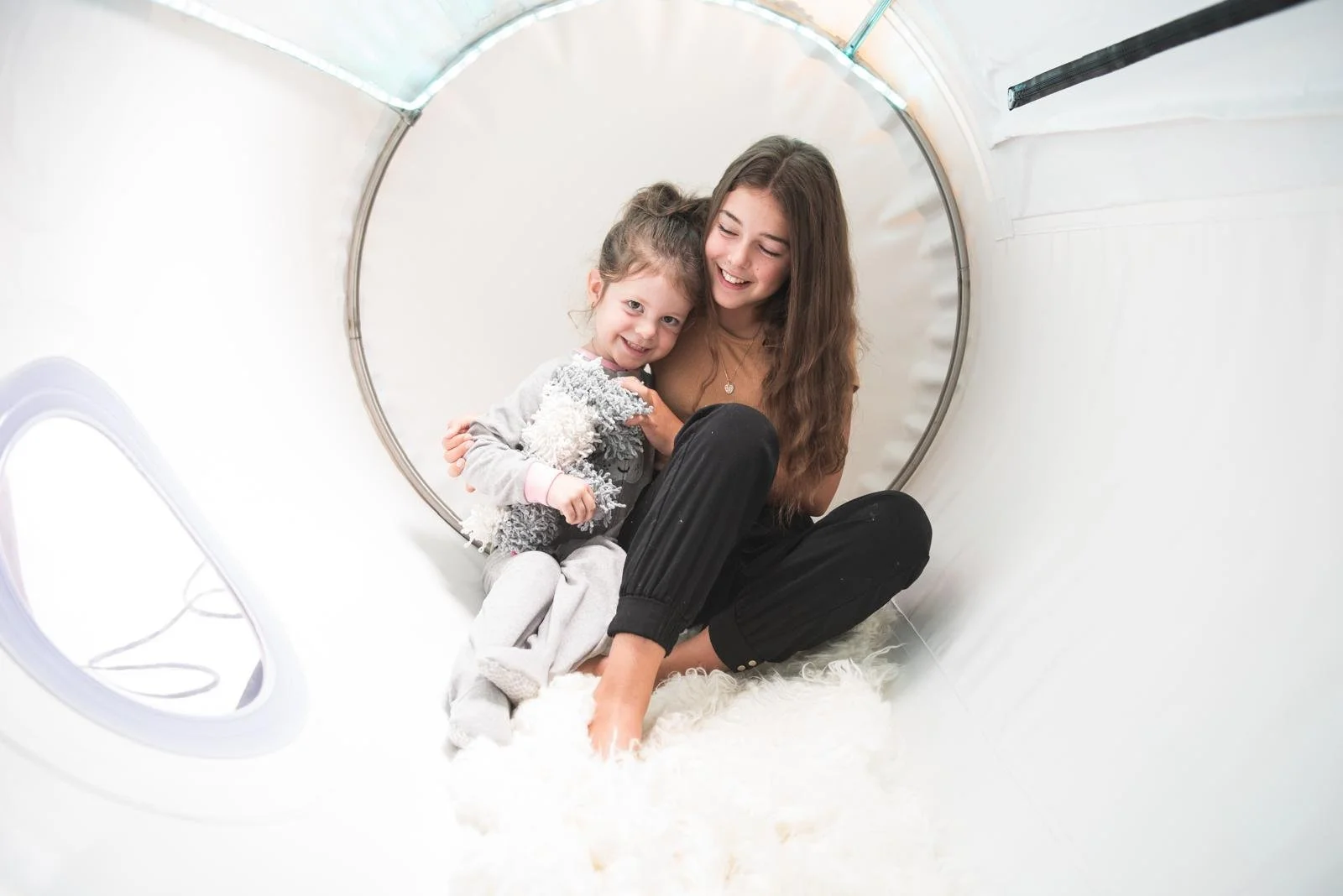Deux filles souriantes, une jeune femme et une enfant, assises à l'intérieur d'un tube de jeu en forme de cœur, entourées de blanc, partageant un moment heureux.