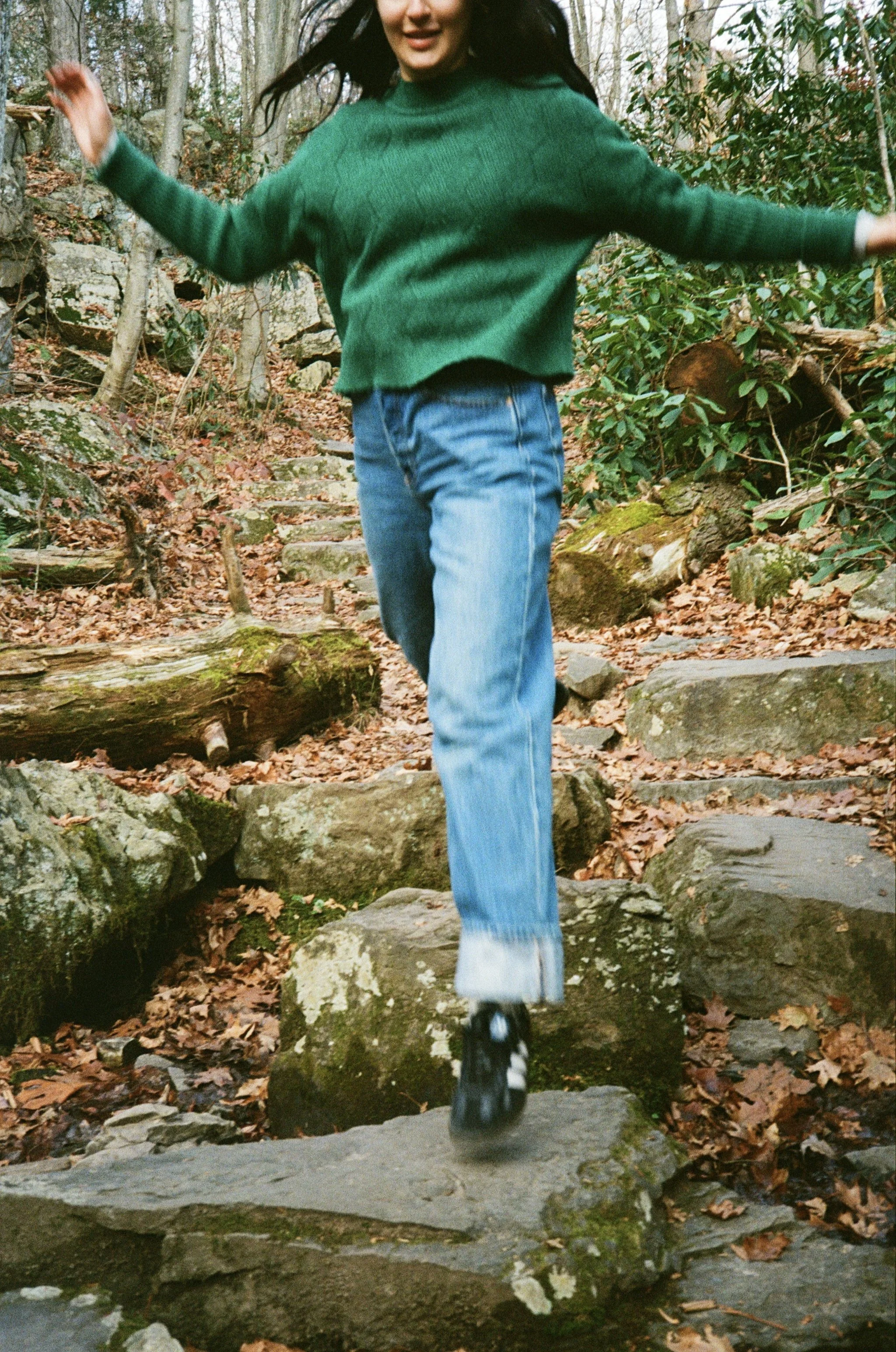 Person in green sweater and jeans walking on rocky trail in wooded area.