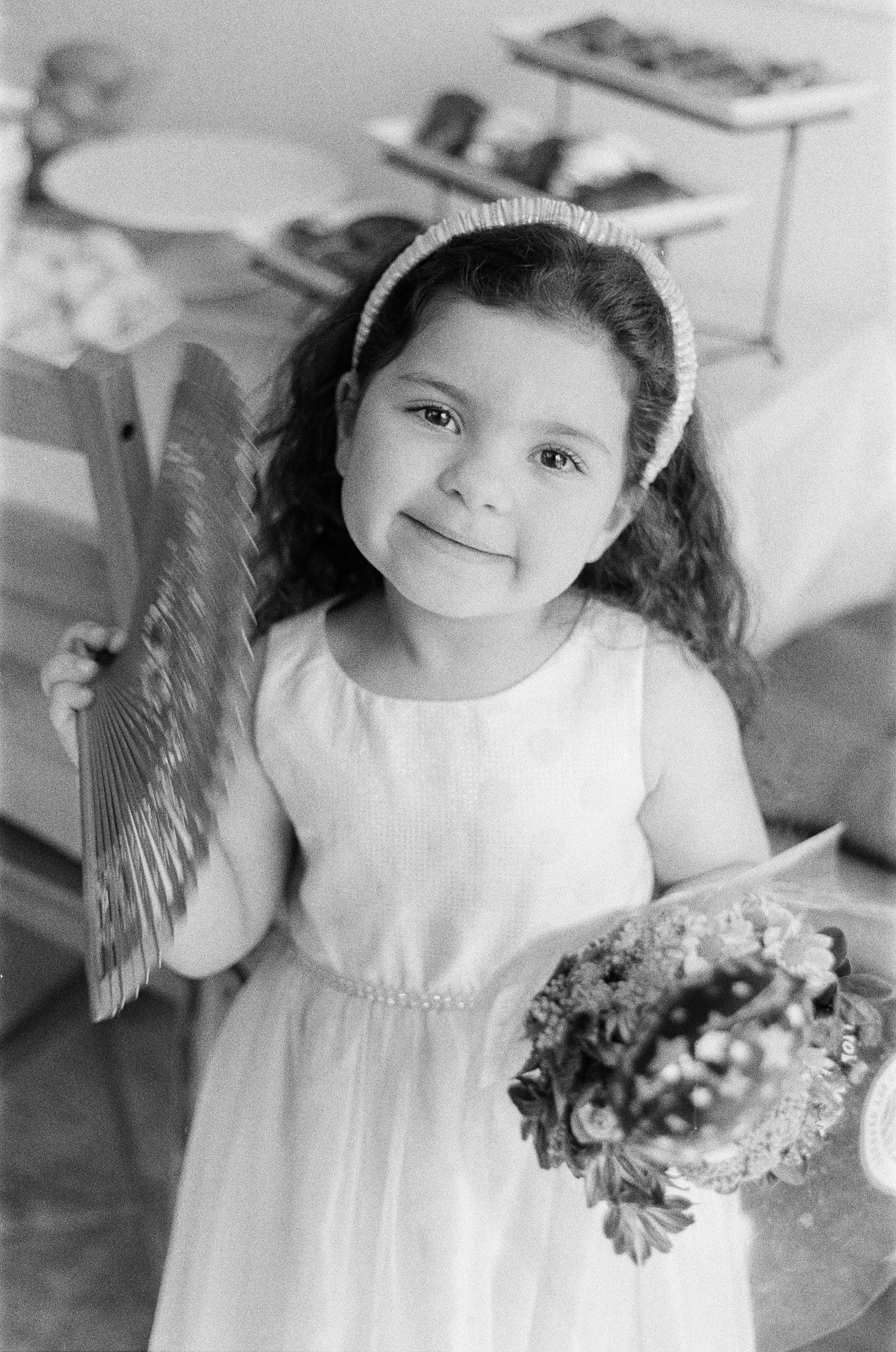 A young girl with curly hair and a headband, wearing a sleeveless dress, holding a fan and a bouquet of flowers, smiling at the camera in a room with shelves in the background.