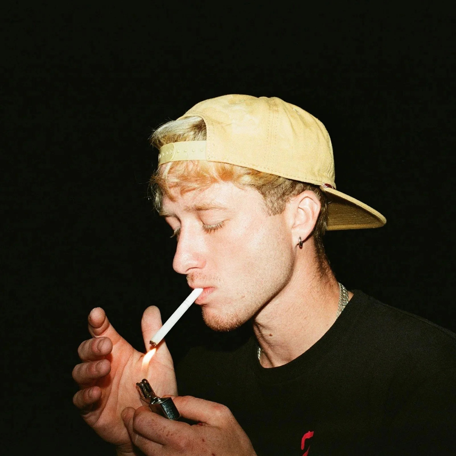 A young man with blonde hair, wearing a beige cap backward, lighting a cigarette with a lighter against a black background.