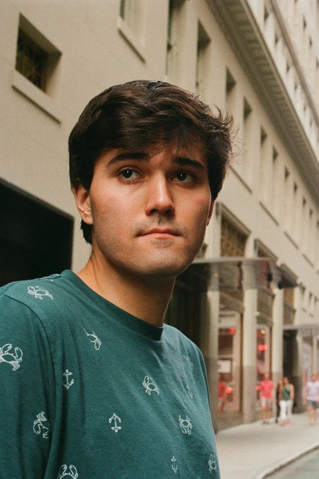 A young man with dark brown hair standing on a city street in front of a beige building with multiple windows. He is wearing a teal T-shirt with nautical symbols and looking slightly to the side.
