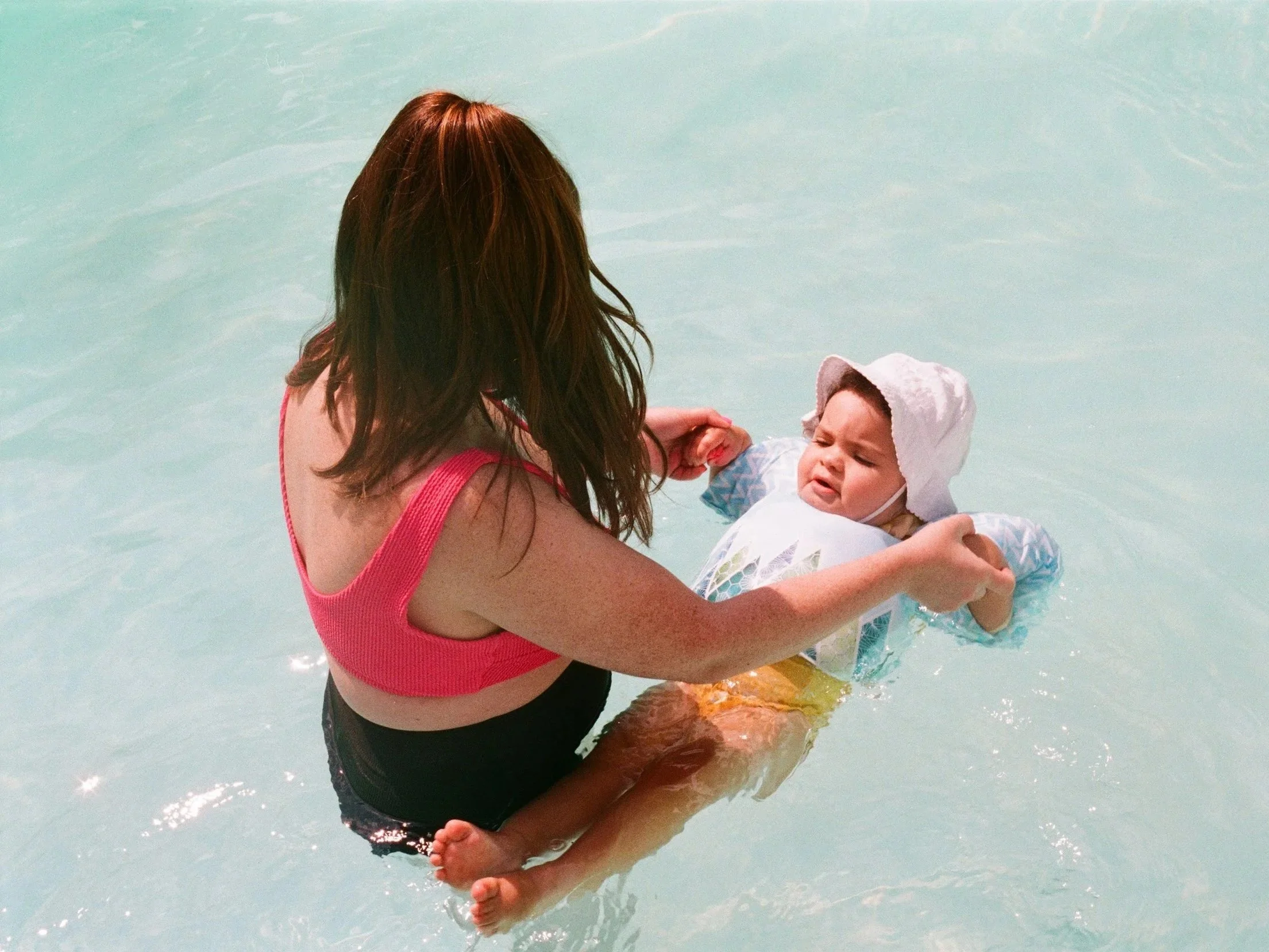 A woman in a pink top holding a crying baby in a swimming pool.