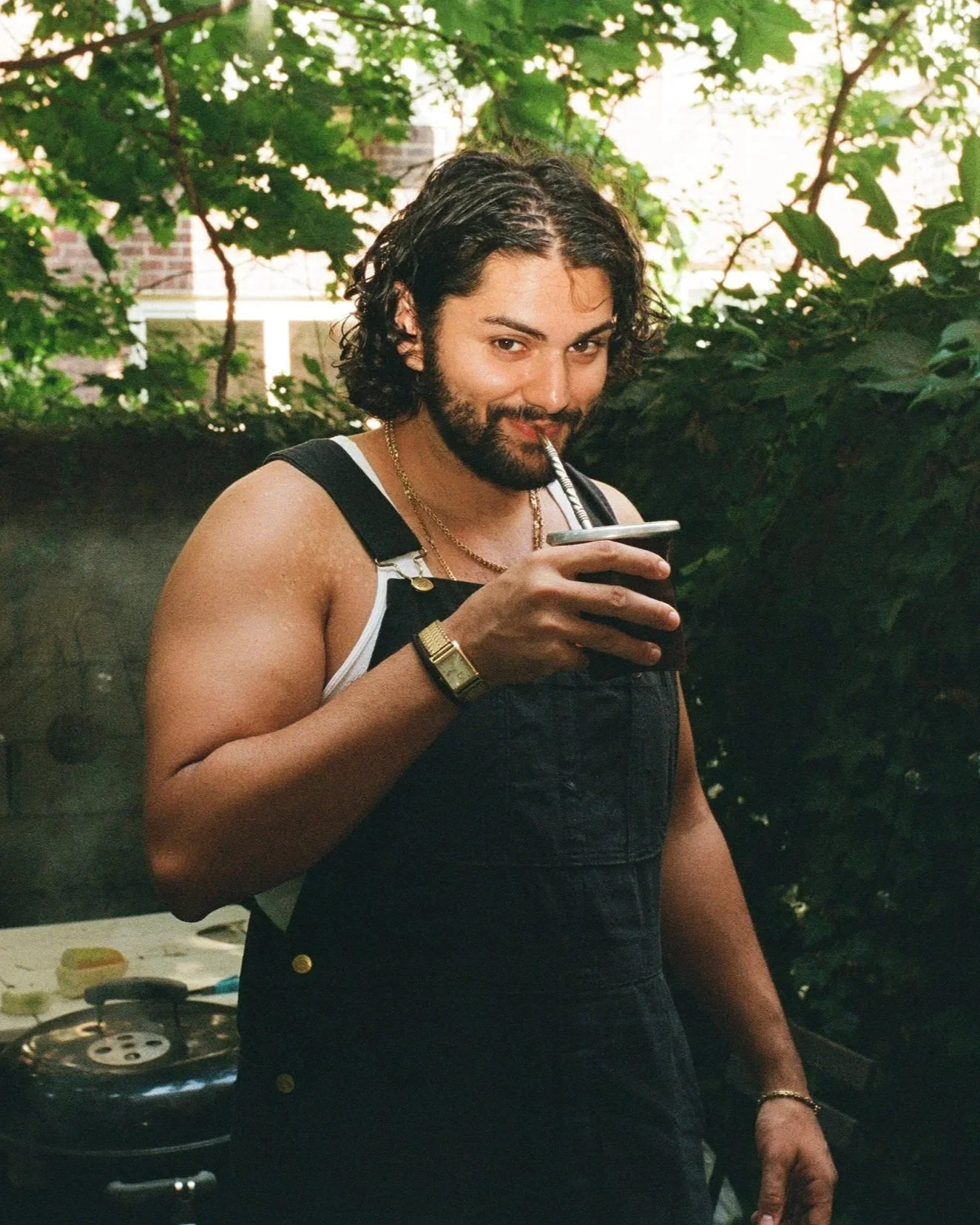 A man with curly hair and a beard, wearing a black sleeveless shirt and gold jewelry, is drinking from a cup with a straw, outdoors with green foliage in the background.