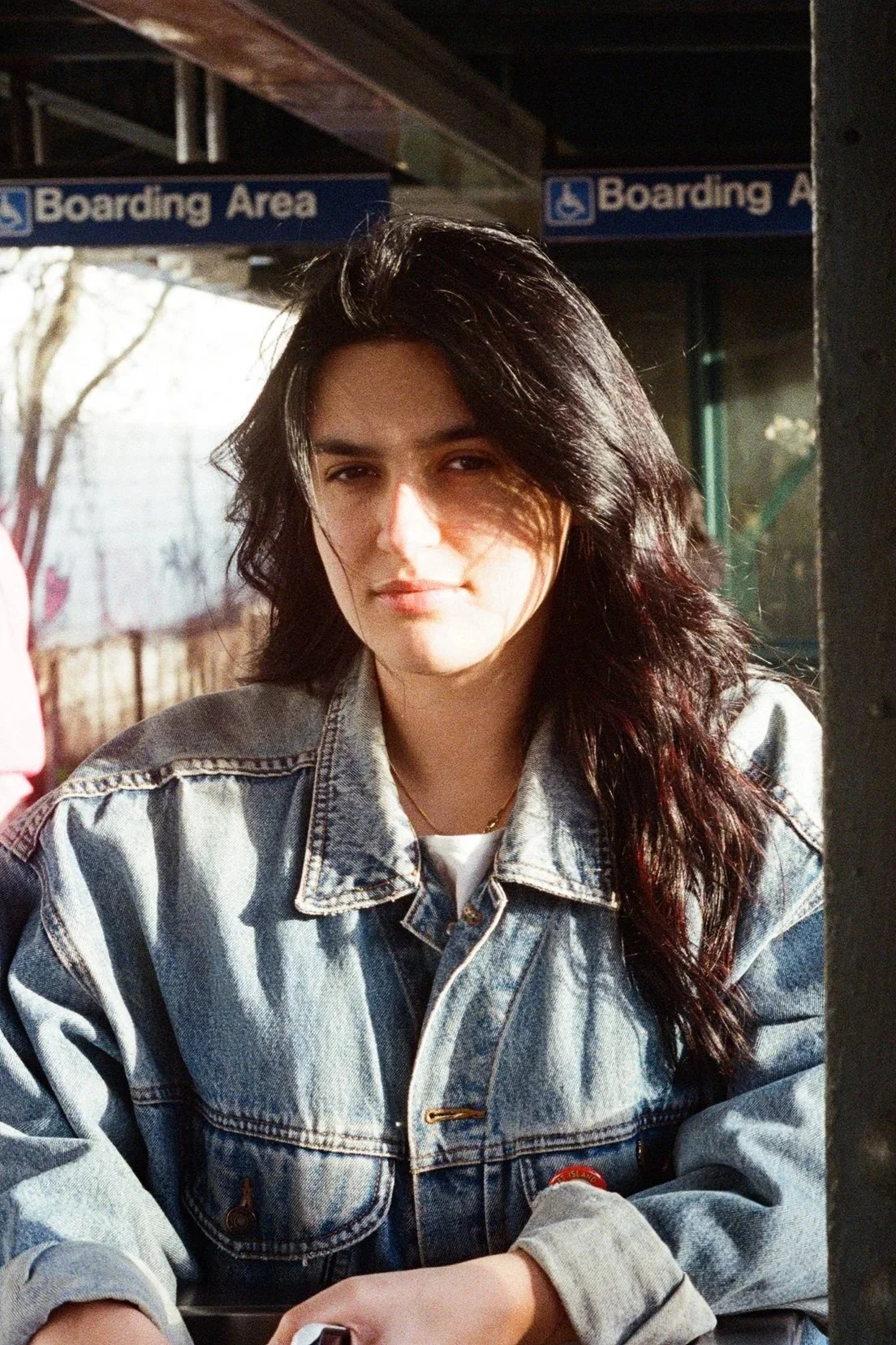 A woman with shoulder-length dark wavy hair wearing a denim jacket sitting at an outdoor location, with the background showing signs that say 'Boarding Area.'