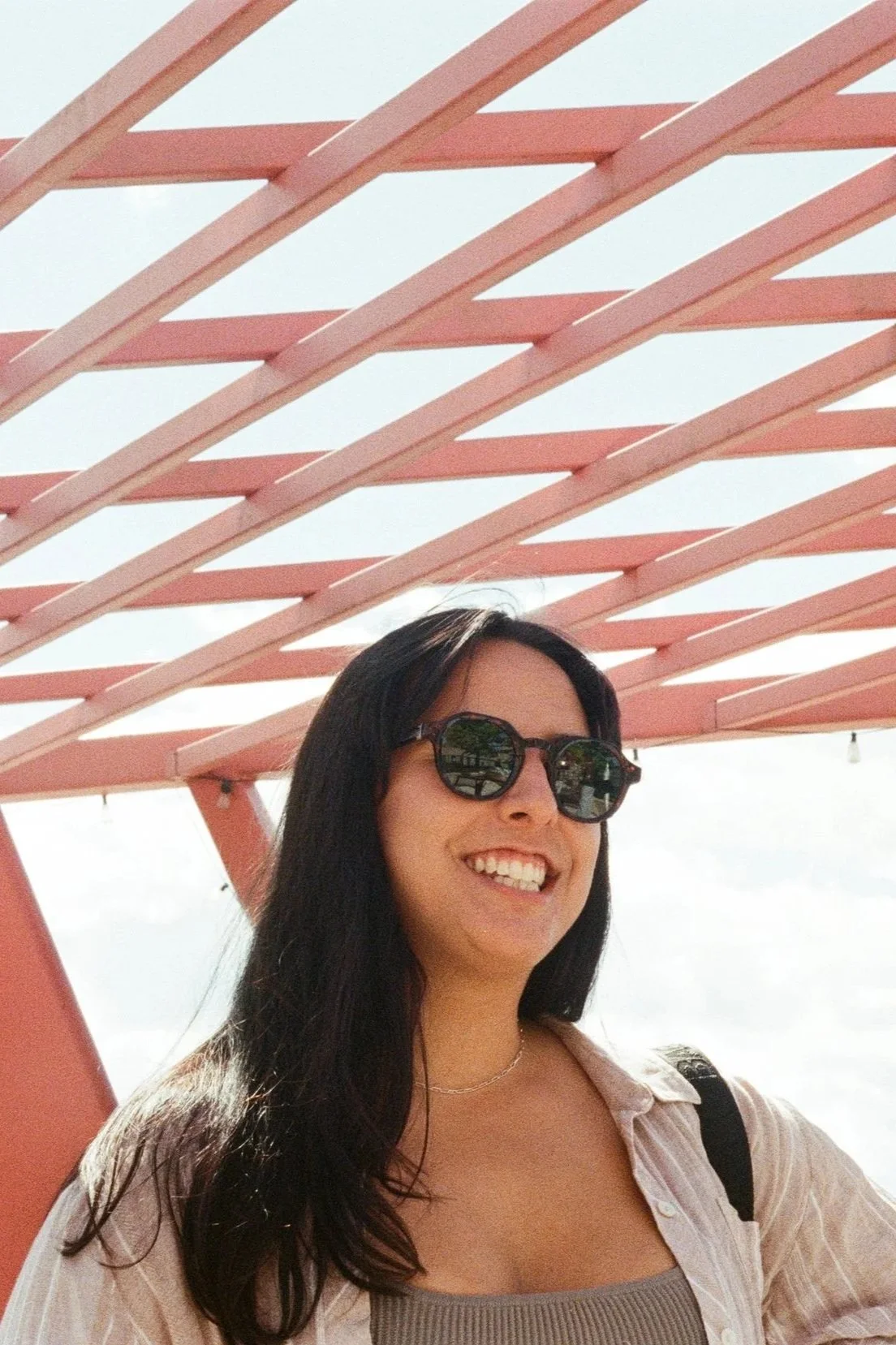 A smiling woman with long black hair wearing sunglasses, a beige jacket, and a gray top, standing outdoors under pink geometric metal structures with a cloudy sky in the background.